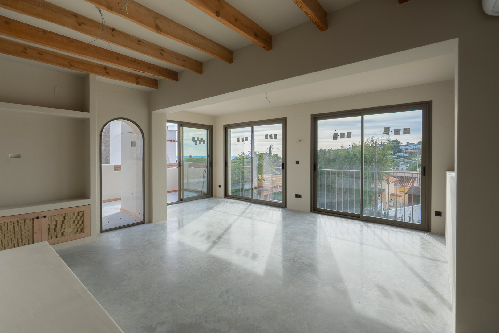 Salon lumineux avec poutres en bois apparentes, sol en béton poli et grandes baies vitrées offrant une vue sur la mer. Étagères intégrées avec finitions en argile naturelle.