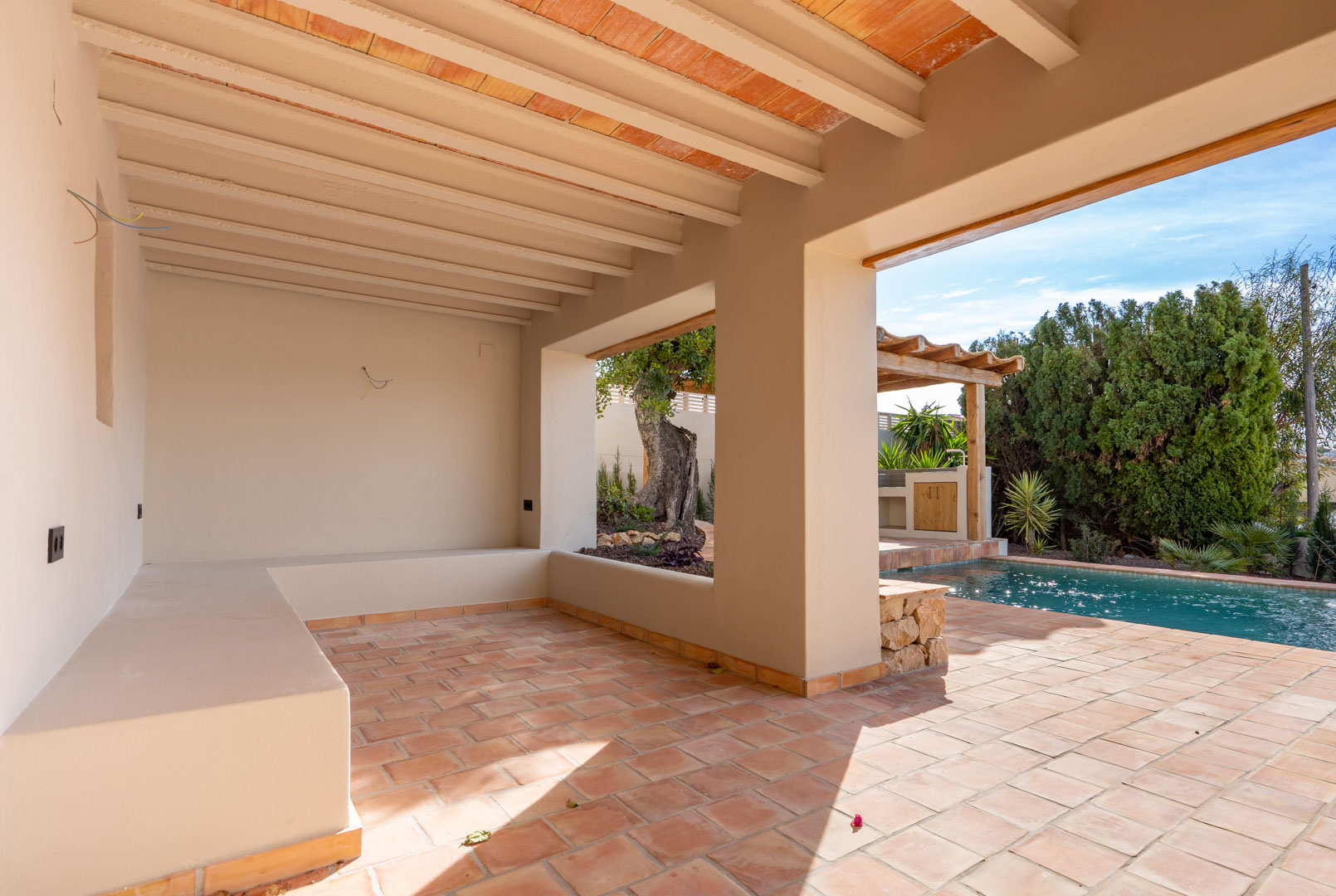 Terrasse couverte avec sols en terre cuite, poutres apparentes et banc maçonné, offrant un accès direct à la piscine et à la pergola.