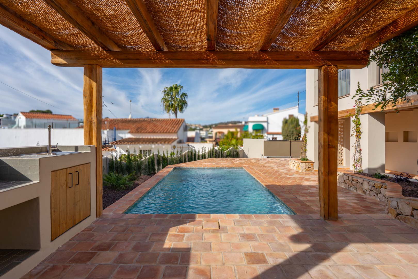 Terrasse extérieure avec sols en terre cuite, pergola en bois et espace barbecue à côté de la piscine nouvellement construite sous le ciel méditerranéen.
