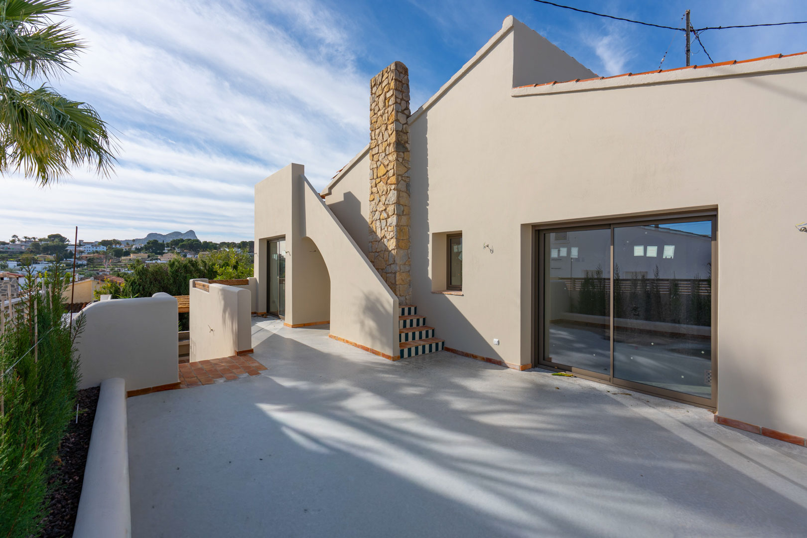 Terrasse supérieure avec sol en béton poli, cheminée en pierre naturelle et vue lointaine sur le célèbre rocher Peñón de Ifach.