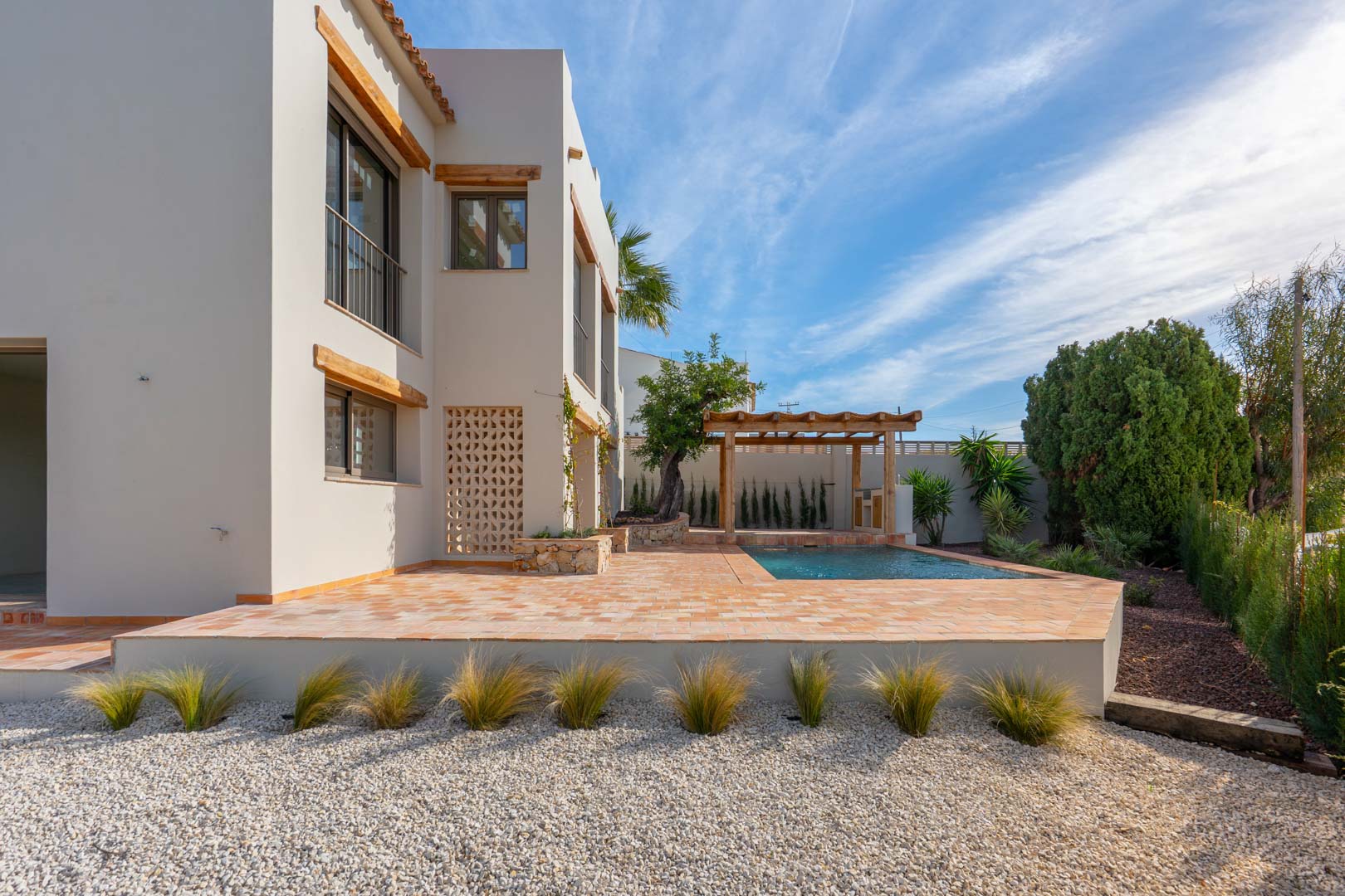 Vue extérieure avec terrasse en terre cuite, piscine neuve et pergola en bois, entourée d'un jardin méditerranéen paysager avec gravier et plantes ornementales.