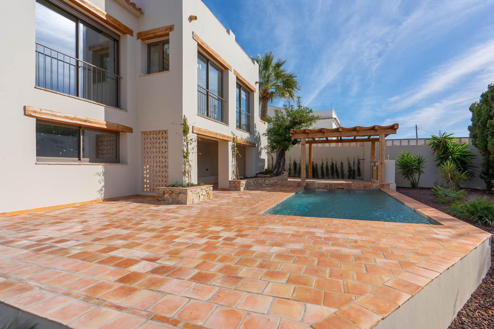 Vue extérieure avec terrasse en terre cuite, piscine privée et pergola en bois. La façade blanche est agrémentée de linteaux en bois et de jardinières en pierre naturelle.