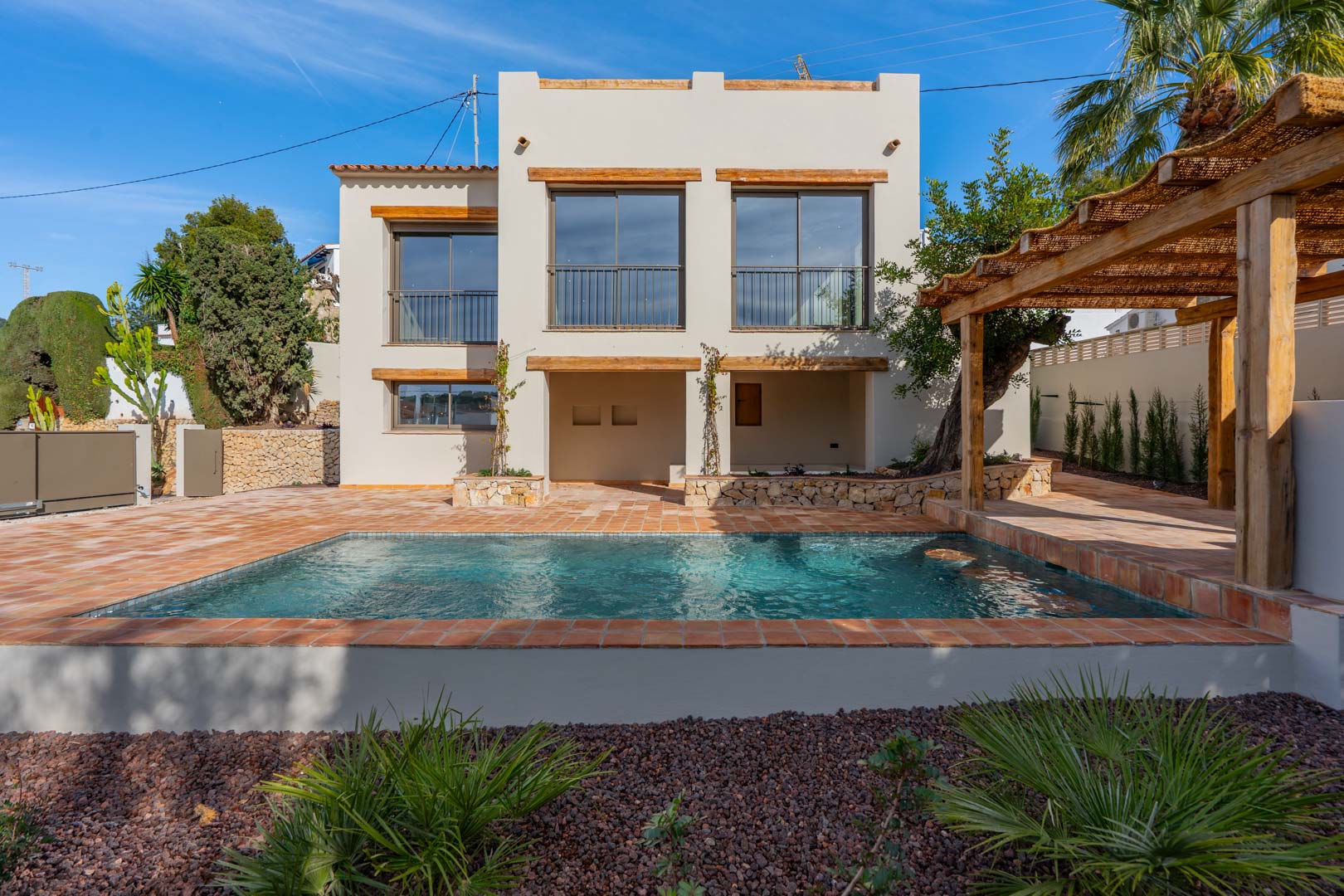 Vue extérieure de la villa avec piscine, terrasse en terre cuite et pergola en bois, mettant en valeur la façade blanche aux linteaux en bois naturel.