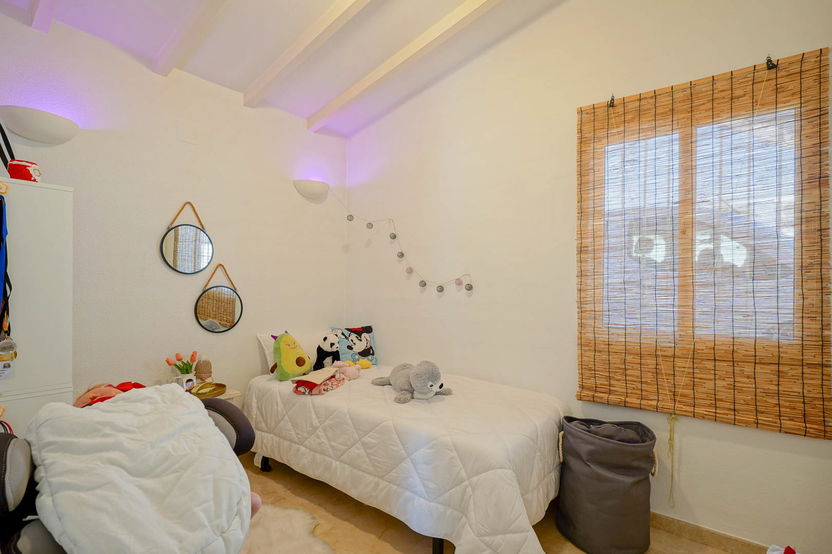 Chambre accueillante avec plafond à poutres blanches, lit simple, miroirs décoratifs suspendus et store en fibres naturelles filtrant la lumière.