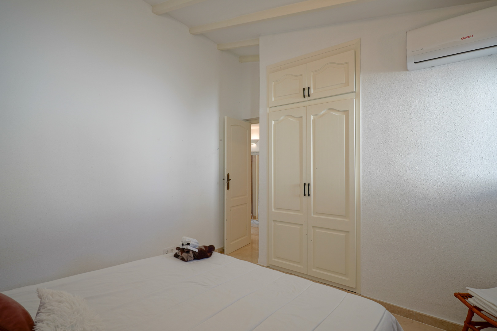 Chambre lumineuse avec armoire encastrée aux portes classiques, poutres apparentes au plafond et climatisation individuelle murale.