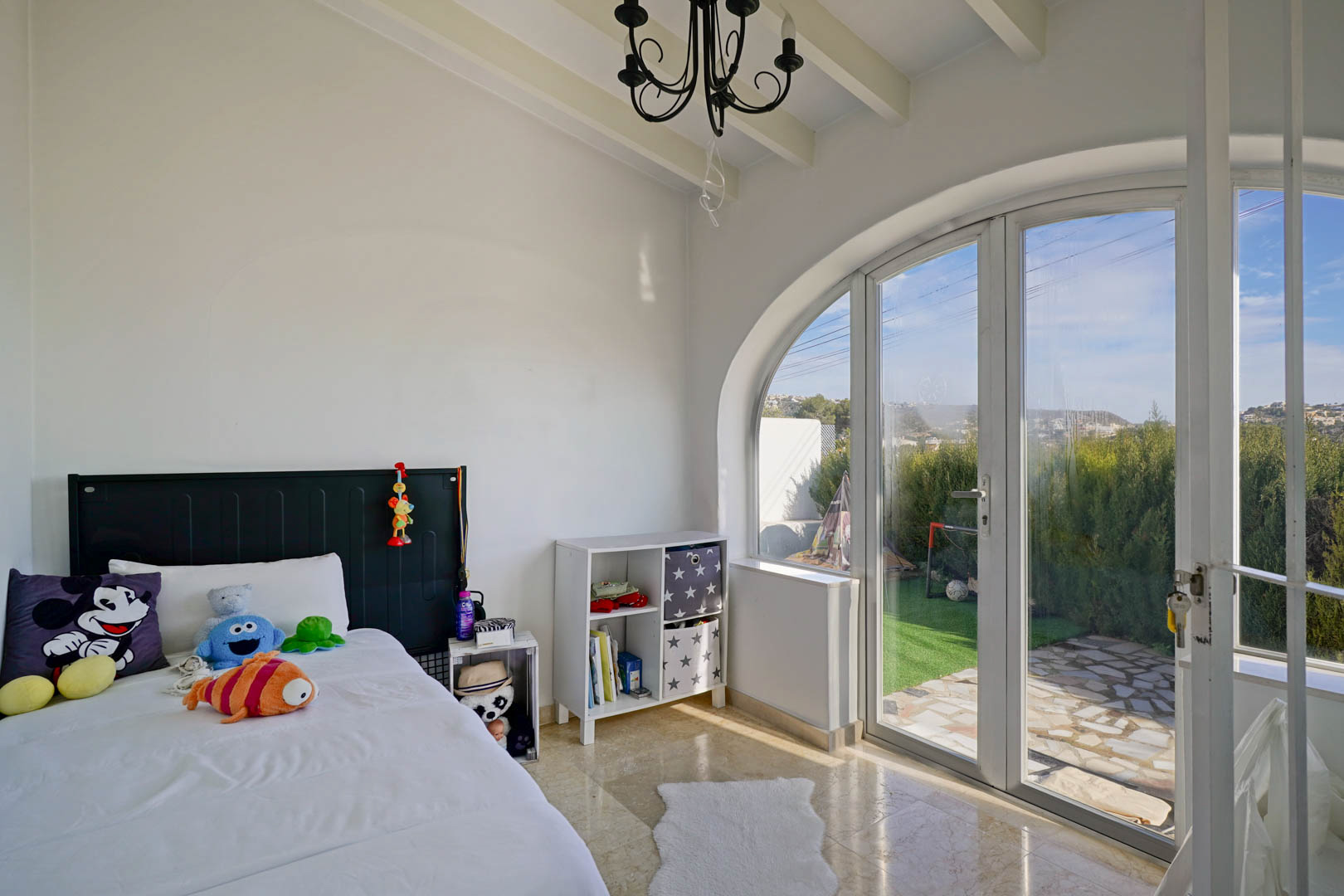 Chambre lumineuse avec plafond à poutres blanches et grande porte vitrée en arche offrant un accès direct au jardin et une belle lumière naturelle.