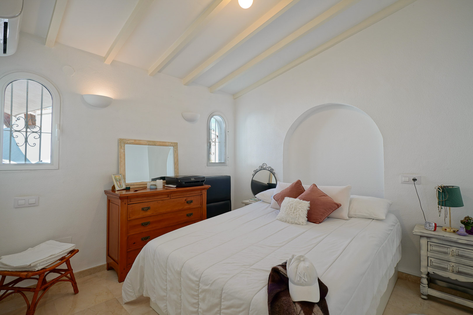 Chambre lumineuse avec poutres apparentes blanches, fenêtres cintrées et niche décorative. Comprend la climatisation et un sol en marbre élégant.
