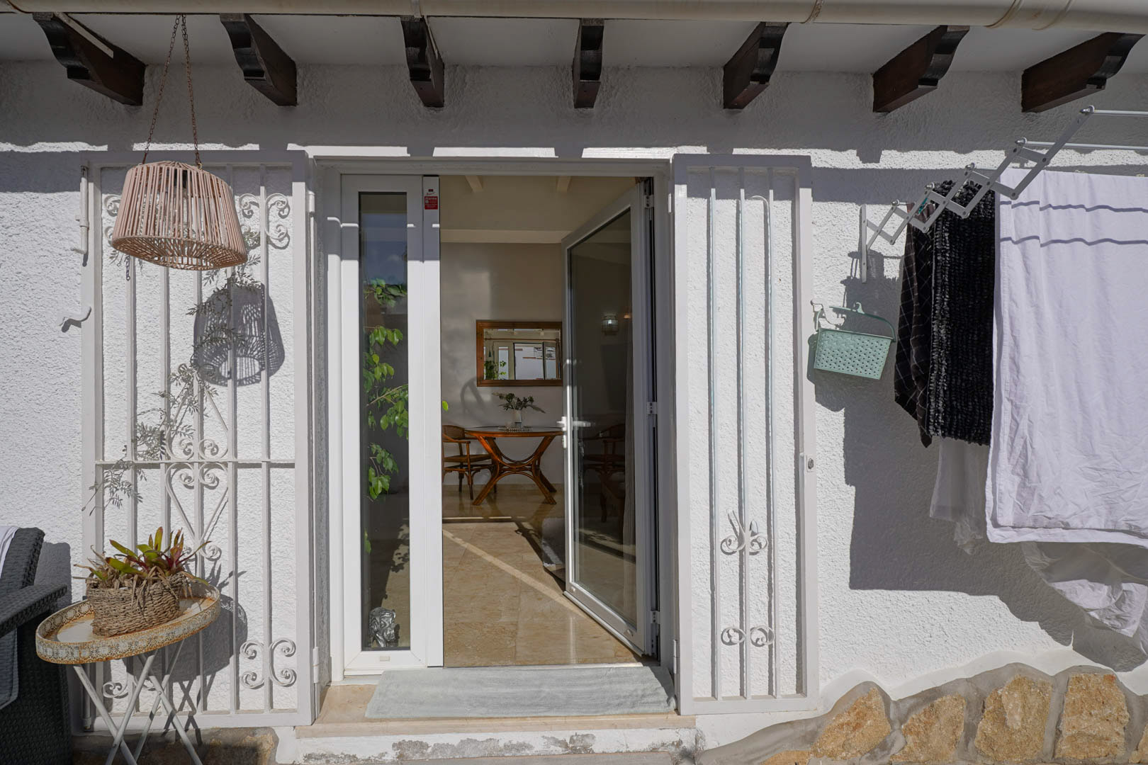 Entrée extérieure avec porte vitrée blanche et grilles de sécurité sous des poutres en bois. On aperçoit le salon intérieur avec son mobilier en bois et son sol en pierre polie.