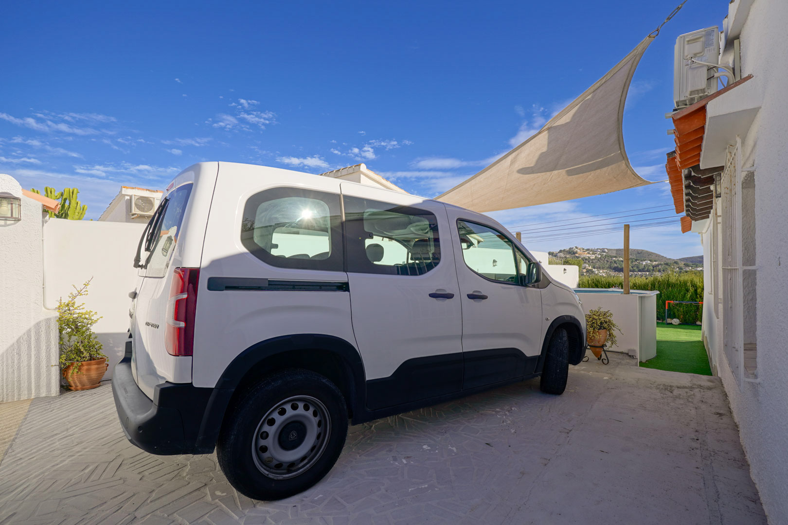 Espace de stationnement privé avec pavage décoratif et voile d'ombrage, situé à côté de la façade blanche de la villa offrant une vue sur le paysage.