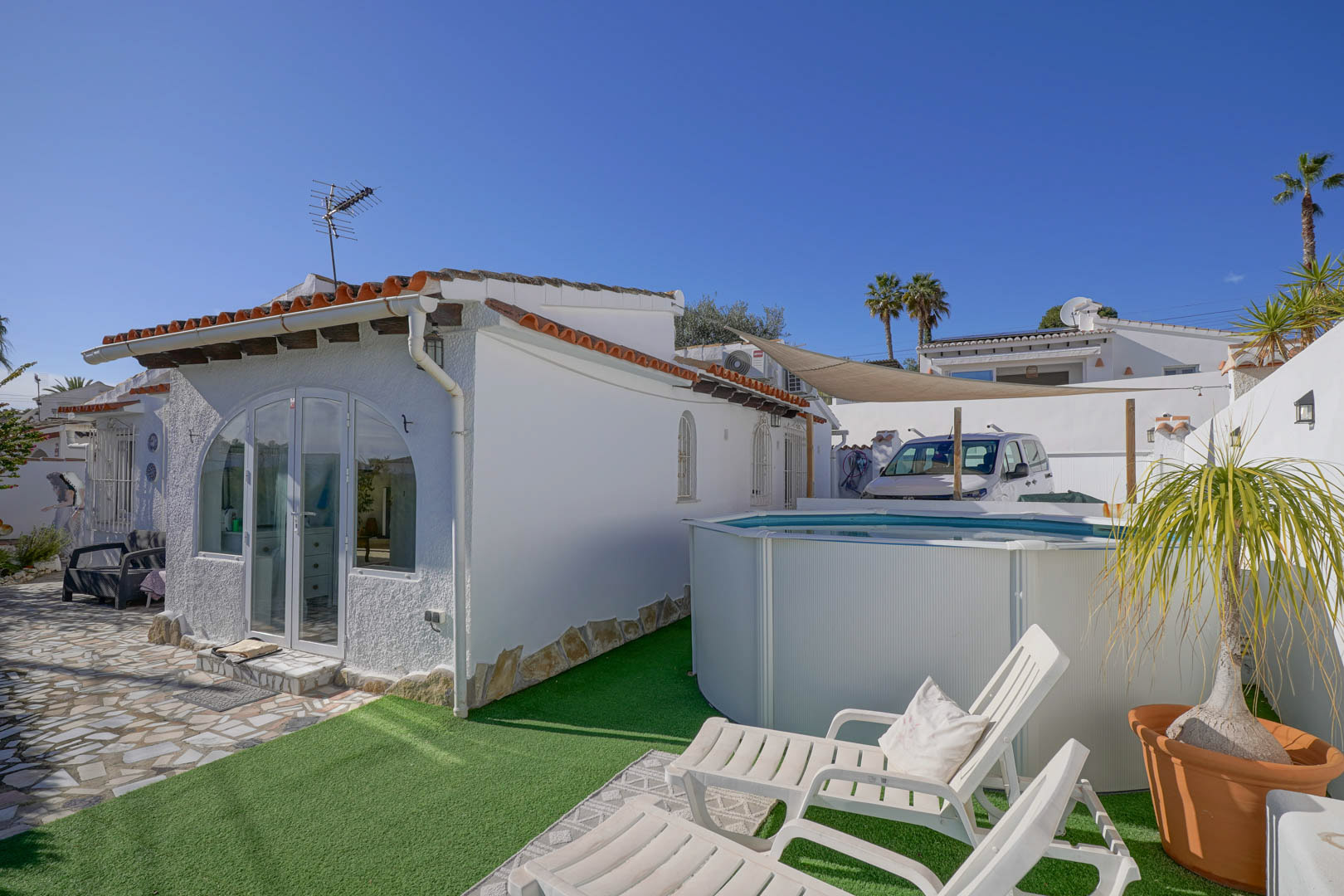 Extérieur de la villa avec façade blanche, porche vitré et espace de détente comprenant une piscine hors-sol sur gazon synthétique et des bains de soleil.