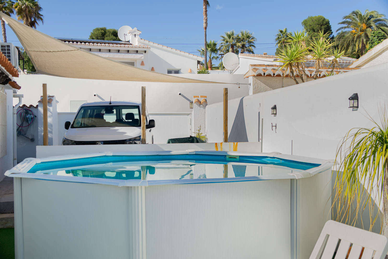 Piscine hors-sol octogonale sur une terrasse ensoleillée avec voile d'ombrage beige, entourée de murs blancs et de palmiers.