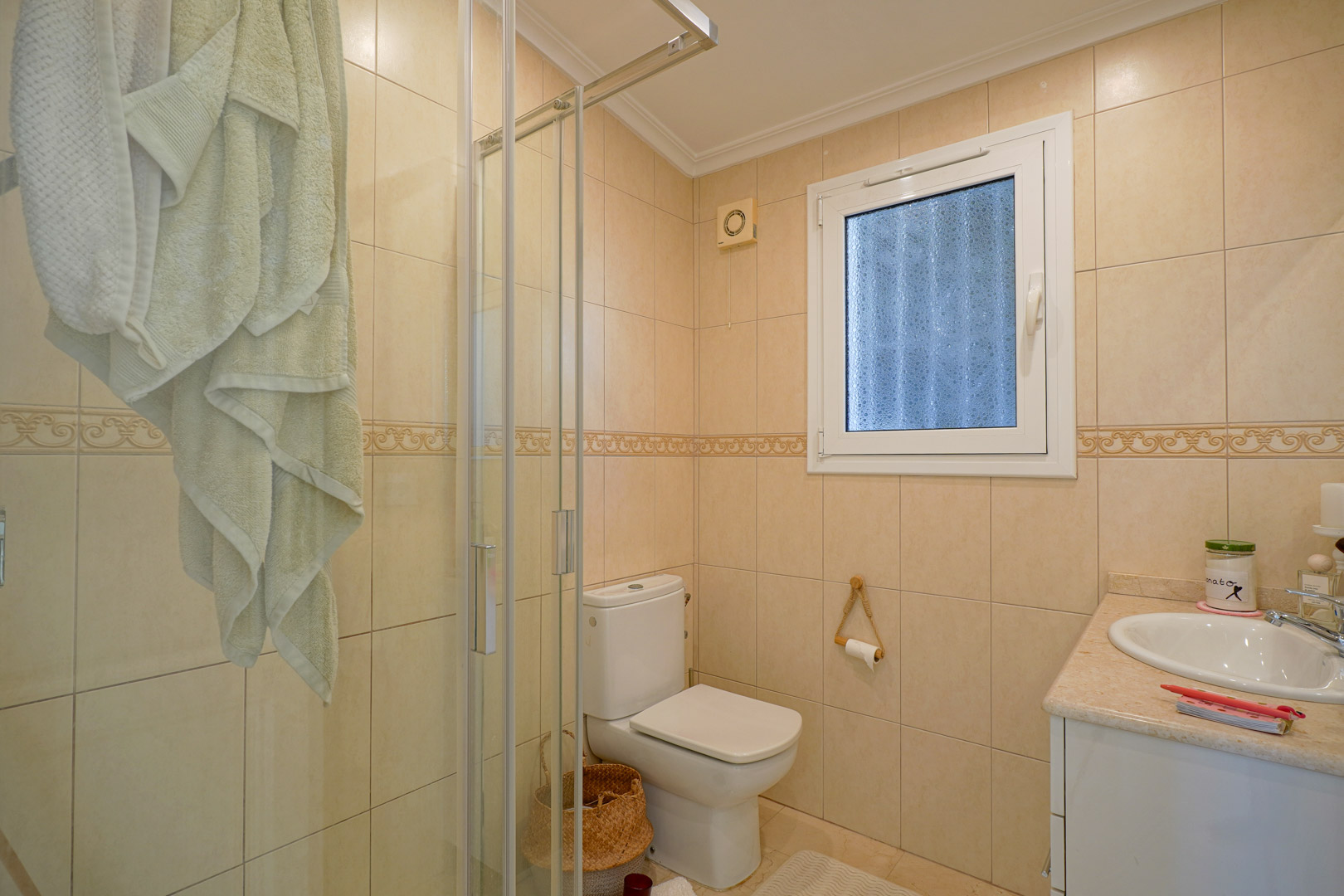 Salle de bain complète avec murs carrelés dans les tons crème, cabine de douche vitrée, sanitaires blancs et meuble vasque avec plan de travail en marbre.