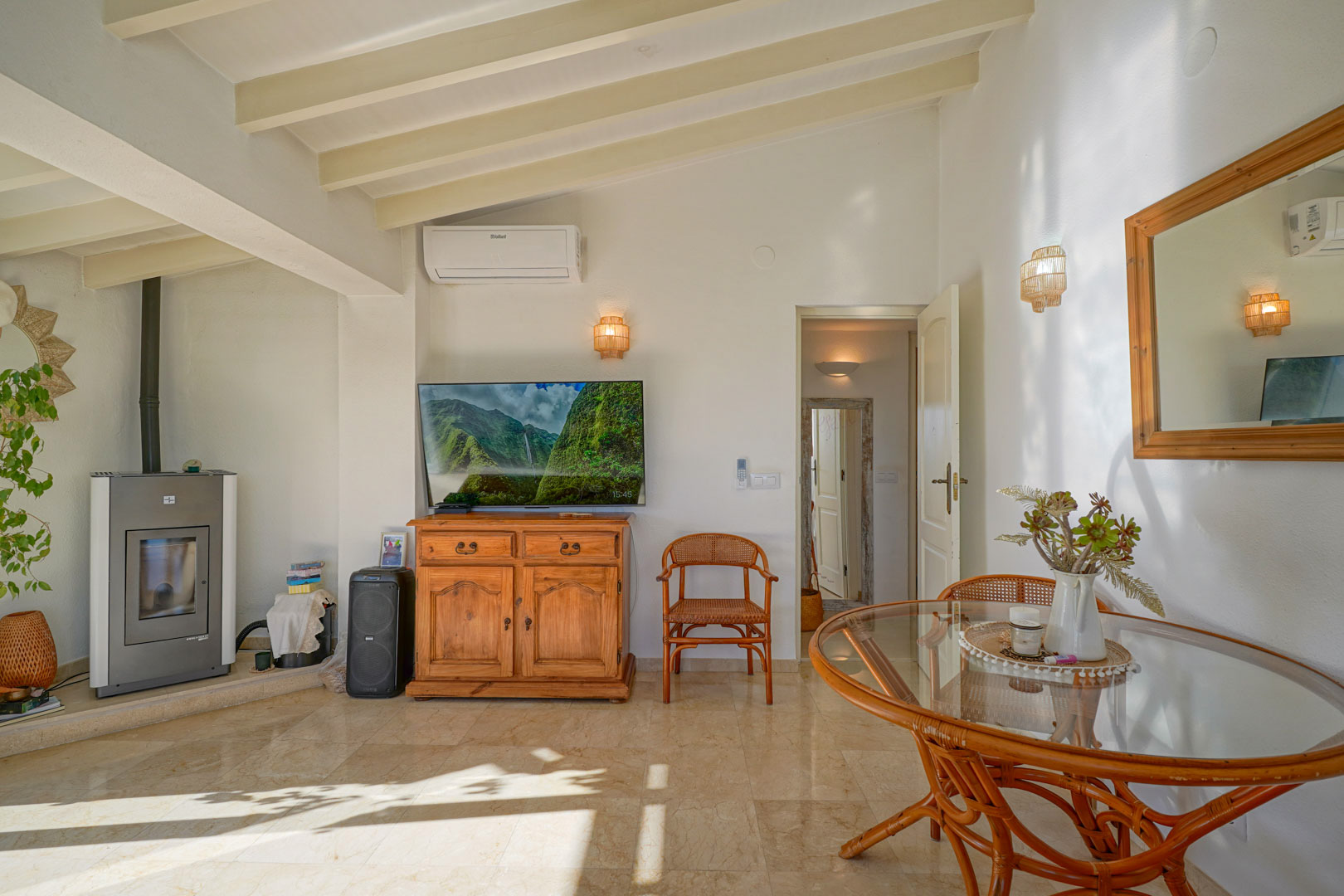 Salon-salle à manger avec poutres apparentes blanches, sol en marbre, poêle à granulés et mobilier en rotin. La pièce dispose de la climatisation et d'une belle lumière naturelle.