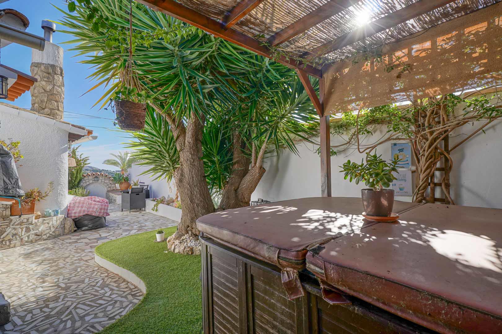 Terrasse extérieure avec jacuzzi couvert sous une pergola en bois, nichée dans un jardin méditerranéen avec des sols en pierre et du gazon synthétique.