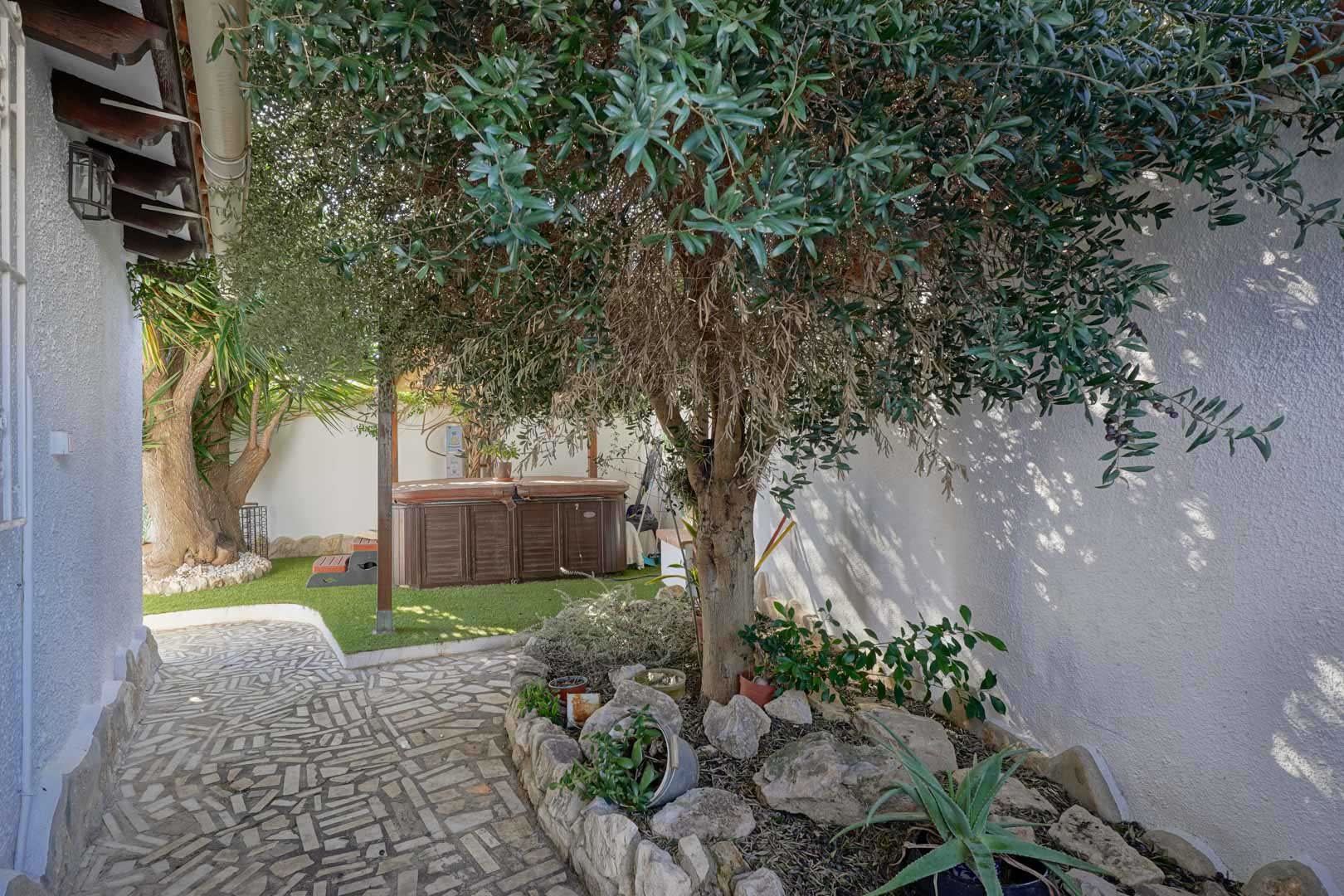 Terrasse extérieure avec jacuzzi privé sur gazon synthétique, agrémentée d'oliviers et d'un chemin en pierre naturelle le long de murs blancs.