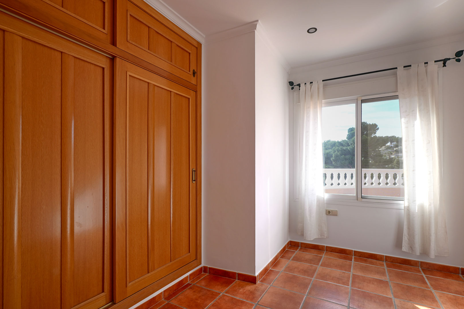 Chambre avec grand placard encastré en bois, sol en terre cuite et fenêtre lumineuse offrant une vue sur la terrasse et la nature environnante.