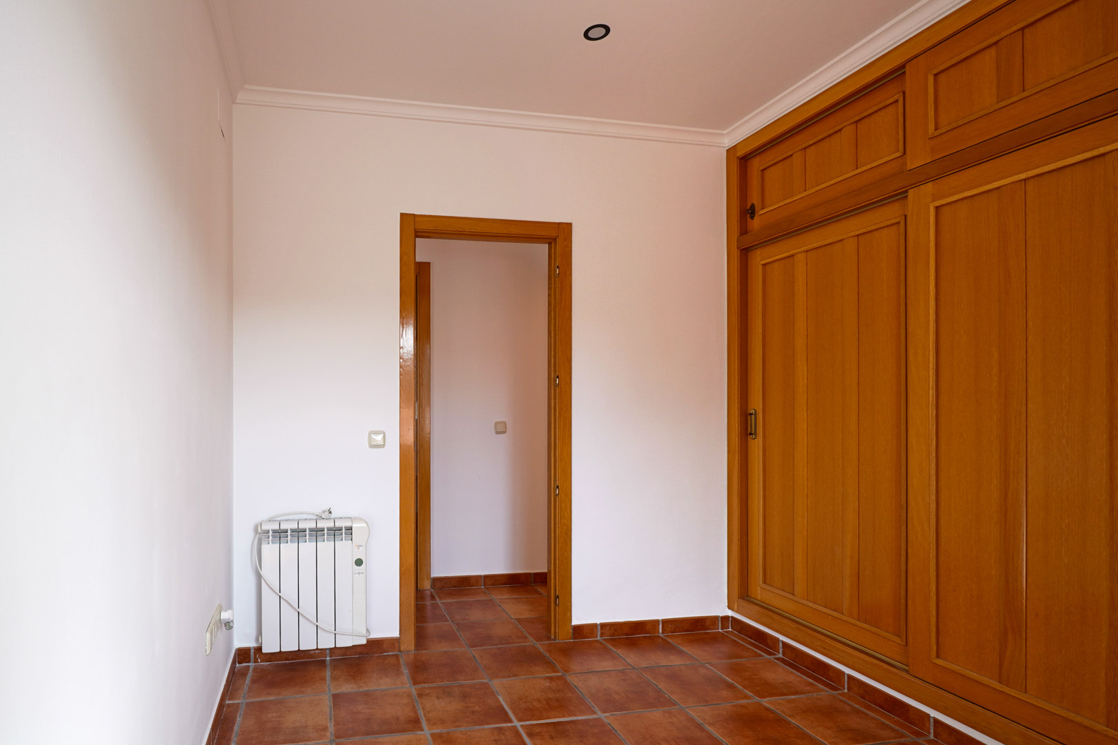 Chambre avec grand placard intégré en bois, sol en grès rustique et radiateur électrique. La pièce dispose de murs blancs, de moulures au plafond et d'un accès au couloir.
