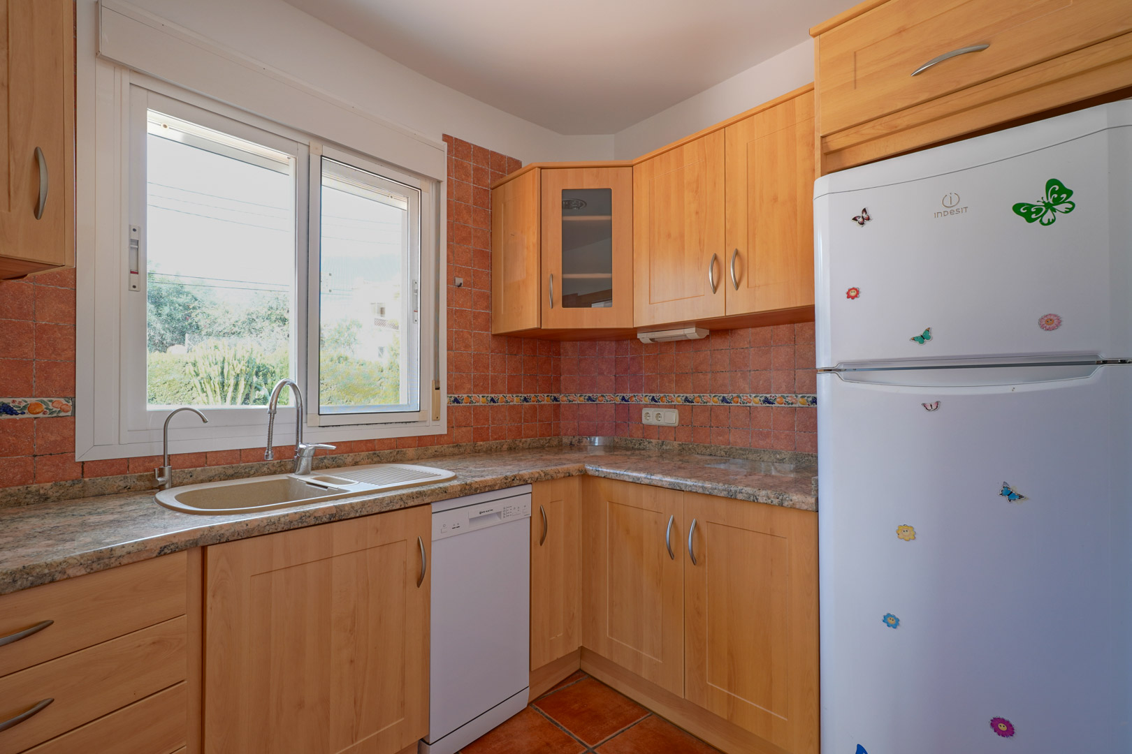 Cuisine indépendante avec meubles en bois clair, plan de travail en granit et carrelage rustique. Équipée d'un lave-vaisselle et d'une fenêtre offrant une luminosité naturelle.