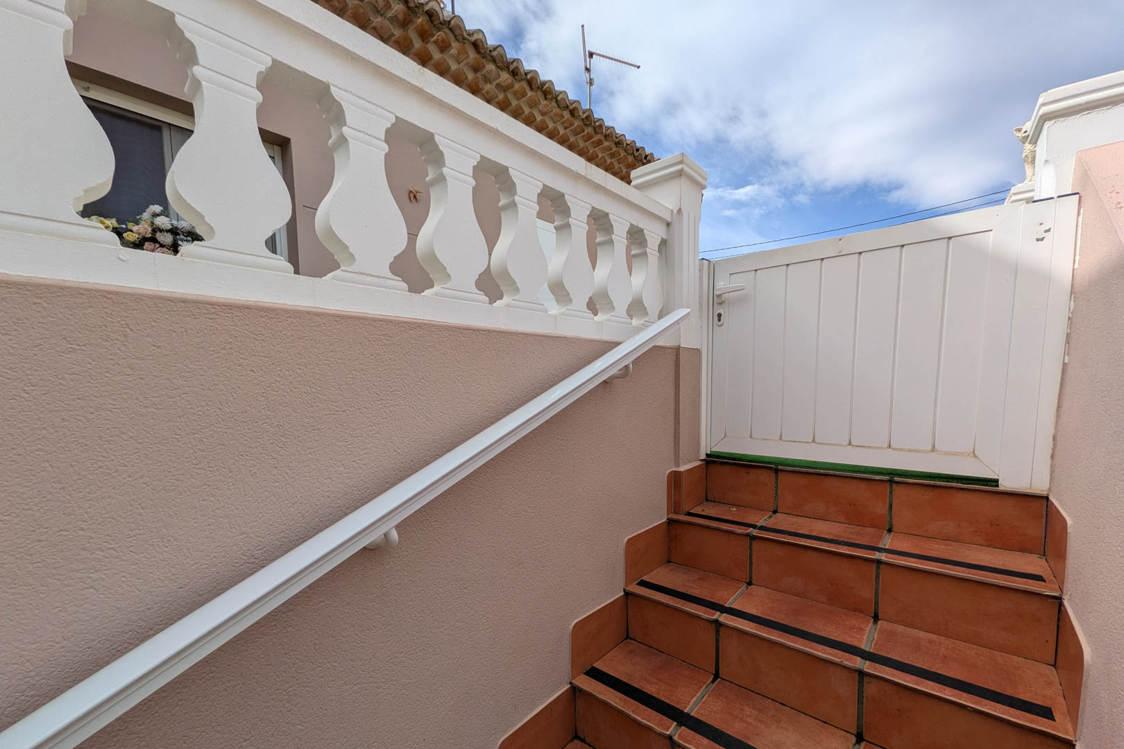Escalier d'accès extérieur avec marches en terre cuite, bandes antidérapantes, main courante blanche et balustrade classique menant au portillon de l'étage.