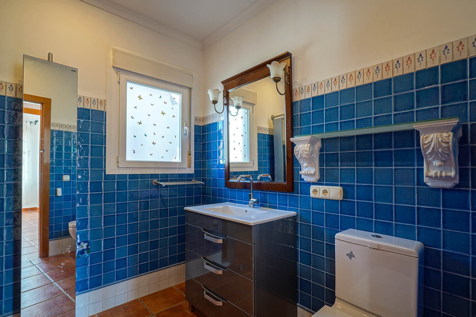 Salle de bain avec carrelage bleu et frise décorative, meuble vasque moderne, miroir à cadre en bois et sol en terre cuite authentique.