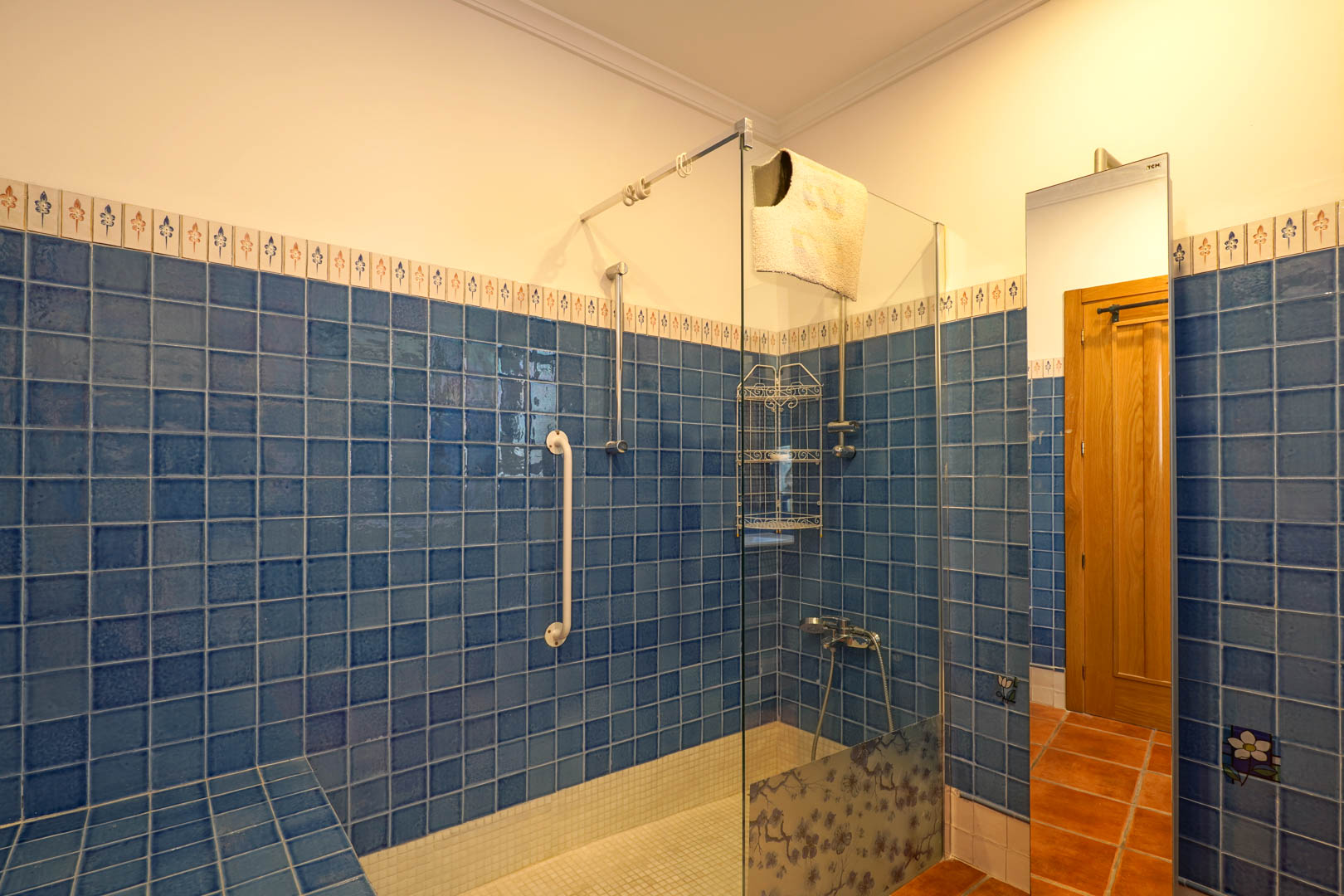 Salle de bain avec douche à l'italienne en carrelage bleu, paroi en verre décorée de motifs floraux, sol en mosaïque blanche et barre d'appui de sécurité.