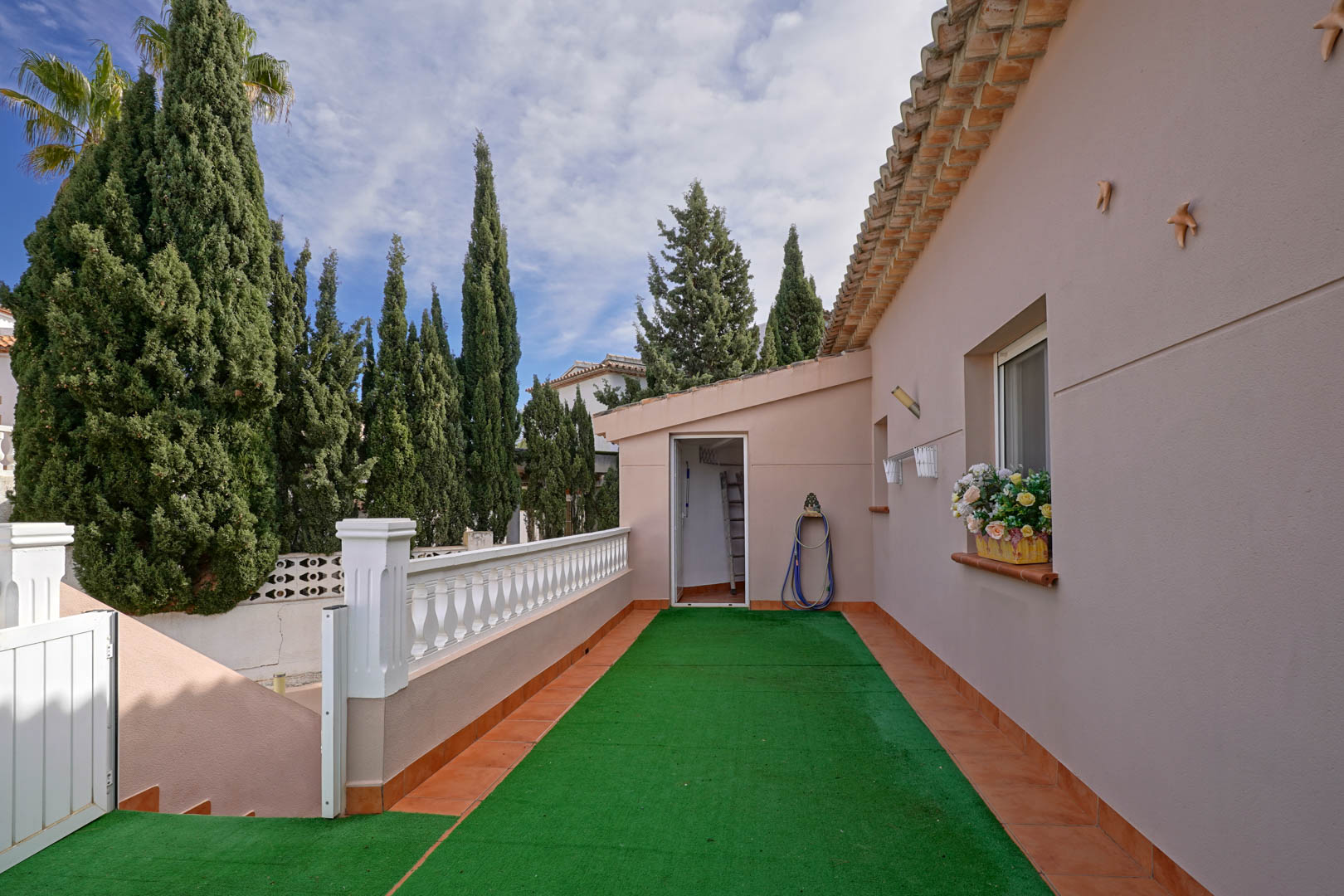 Terrasse d'accès avec gazon artificiel et balustrade blanche, reliant l'entrée principale à la buanderie dans un cadre arboré de cyprès.