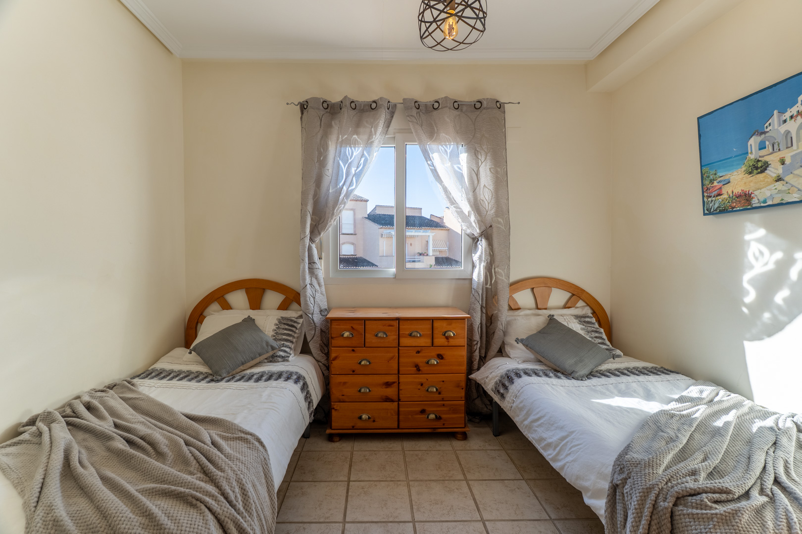 Chambre avec deux lits simples, têtes de lit en bois et commode centrale. Elle dispose de sols en grès, de lumière naturelle et d'un luminaire suspendu moderne.