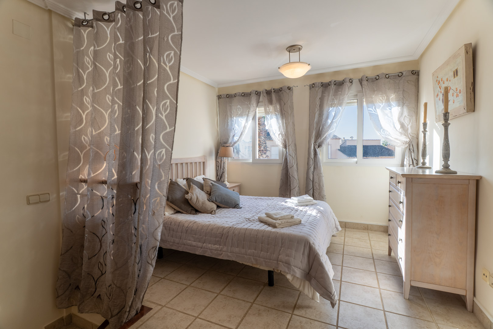 Chambre avec sol en grès, lit double, commode en bois et fenêtres avec rideaux à motifs offrant une luminosité naturelle constante.