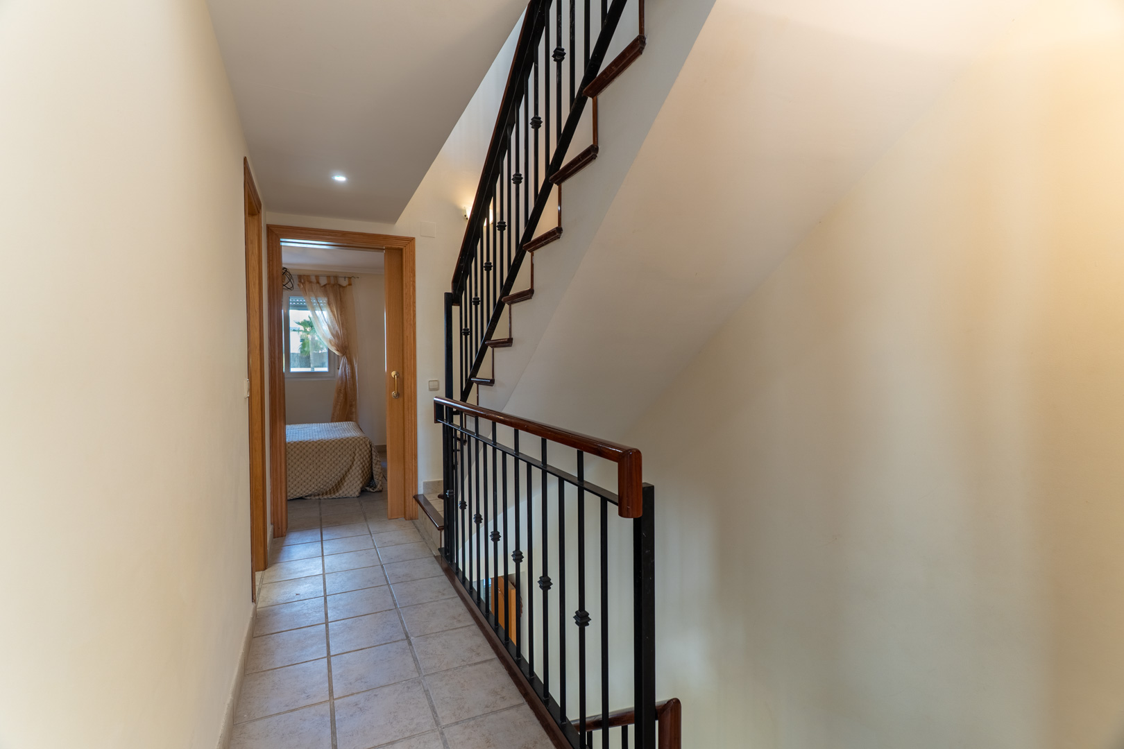 Couloir de distribution avec sol en céramique menant aux chambres, doté d'un escalier avec main courante en bois et ferronnerie noire.