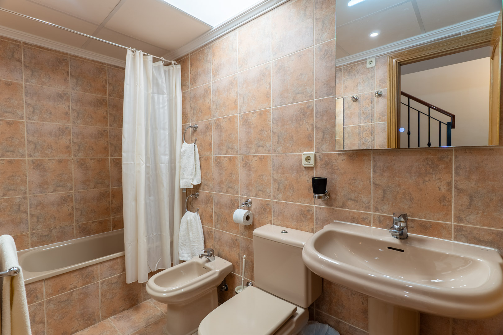 Salle de bain complète avec baignoire, bidet, lavabo sur colonne et carrelage en céramique beige, bénéficiant d'un éclairage zénithal.