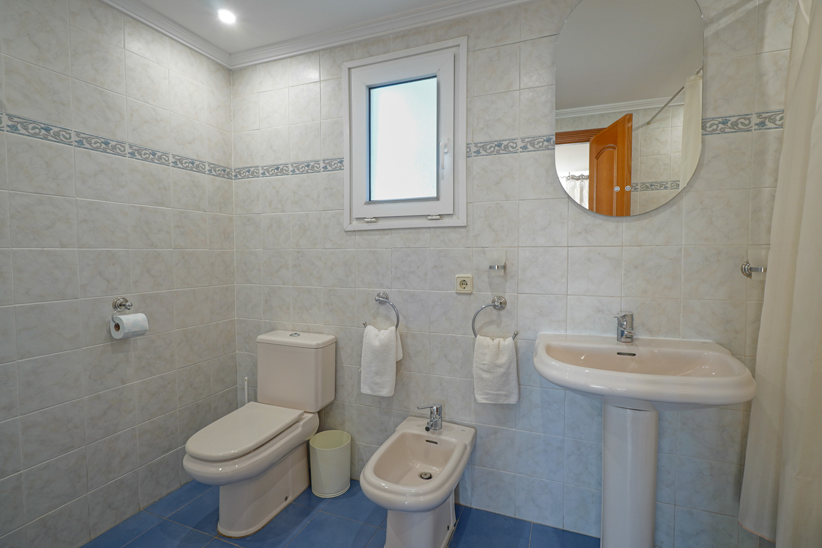 Salle de bain complète avec carrelage clair, frise décorative et sol bleu. Équipée d'un WC, d'un bidet, d'un lavabo sur colonne et d'une fenêtre dépolie.
