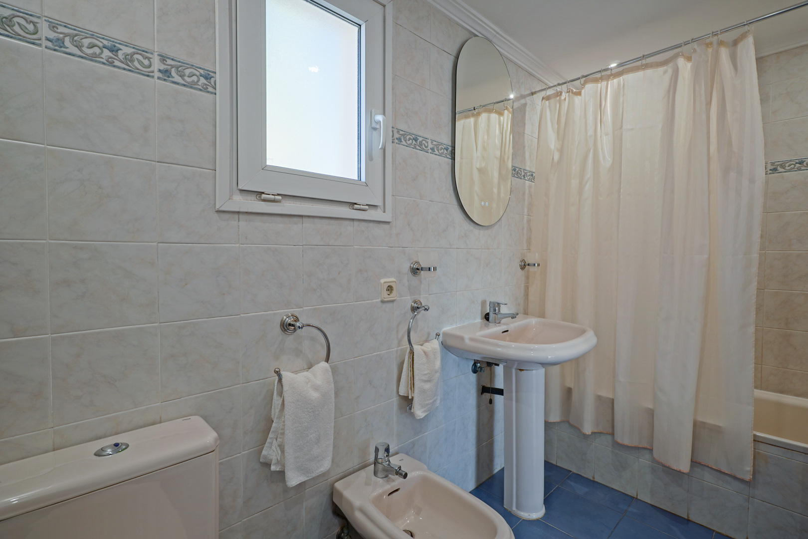 Salle de bain familiale avec sanitaires de couleur crème, bidet et lavabo sur colonne. Elle dispose d'une baignoire, d'un carrelage mural avec frise et d'un sol en grès bleu.