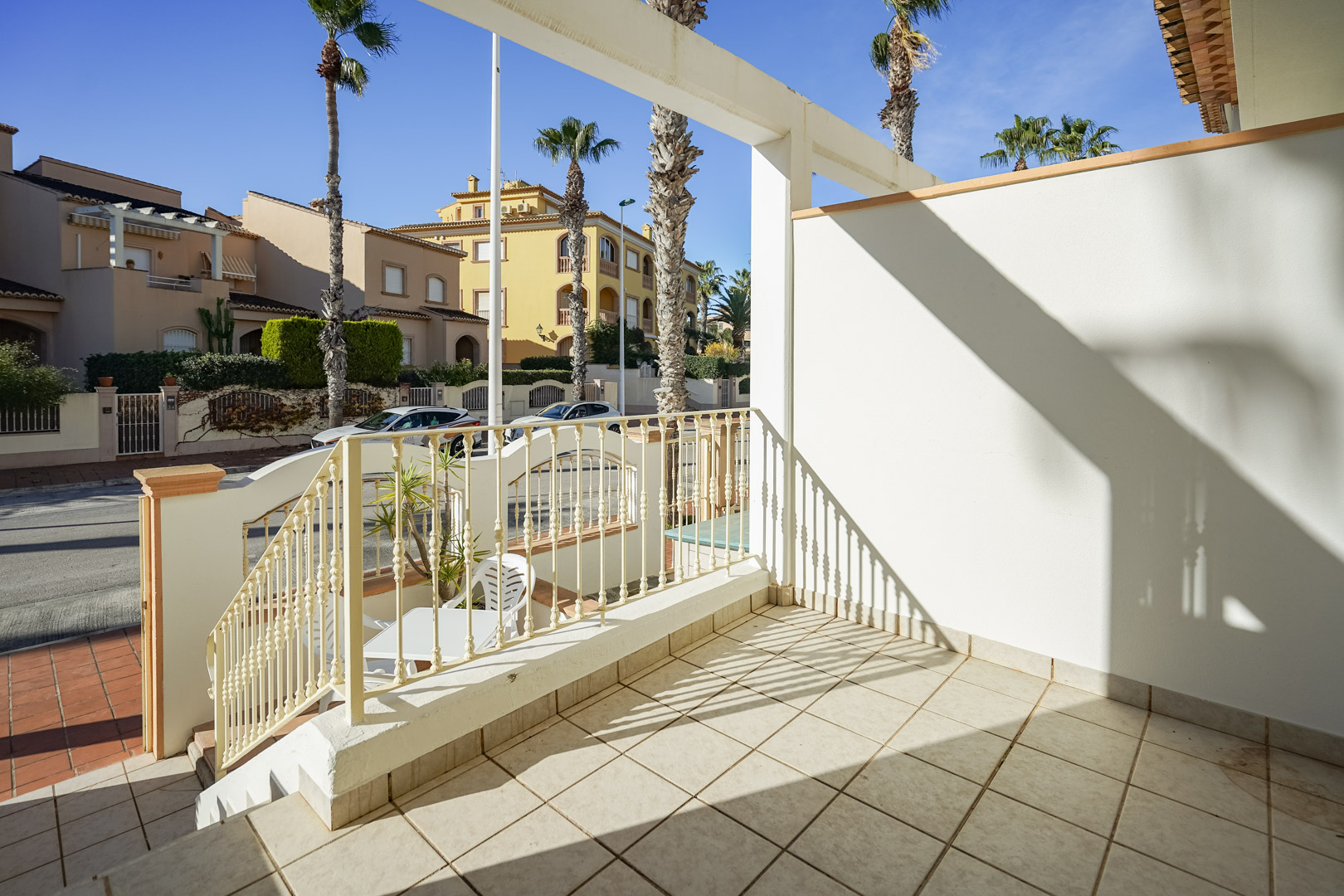 Terrasse avant surélevée avec sol en céramique et garde-corps métallique, offrant une vue sur les palmiers et le quartier résidentiel de Jávea.