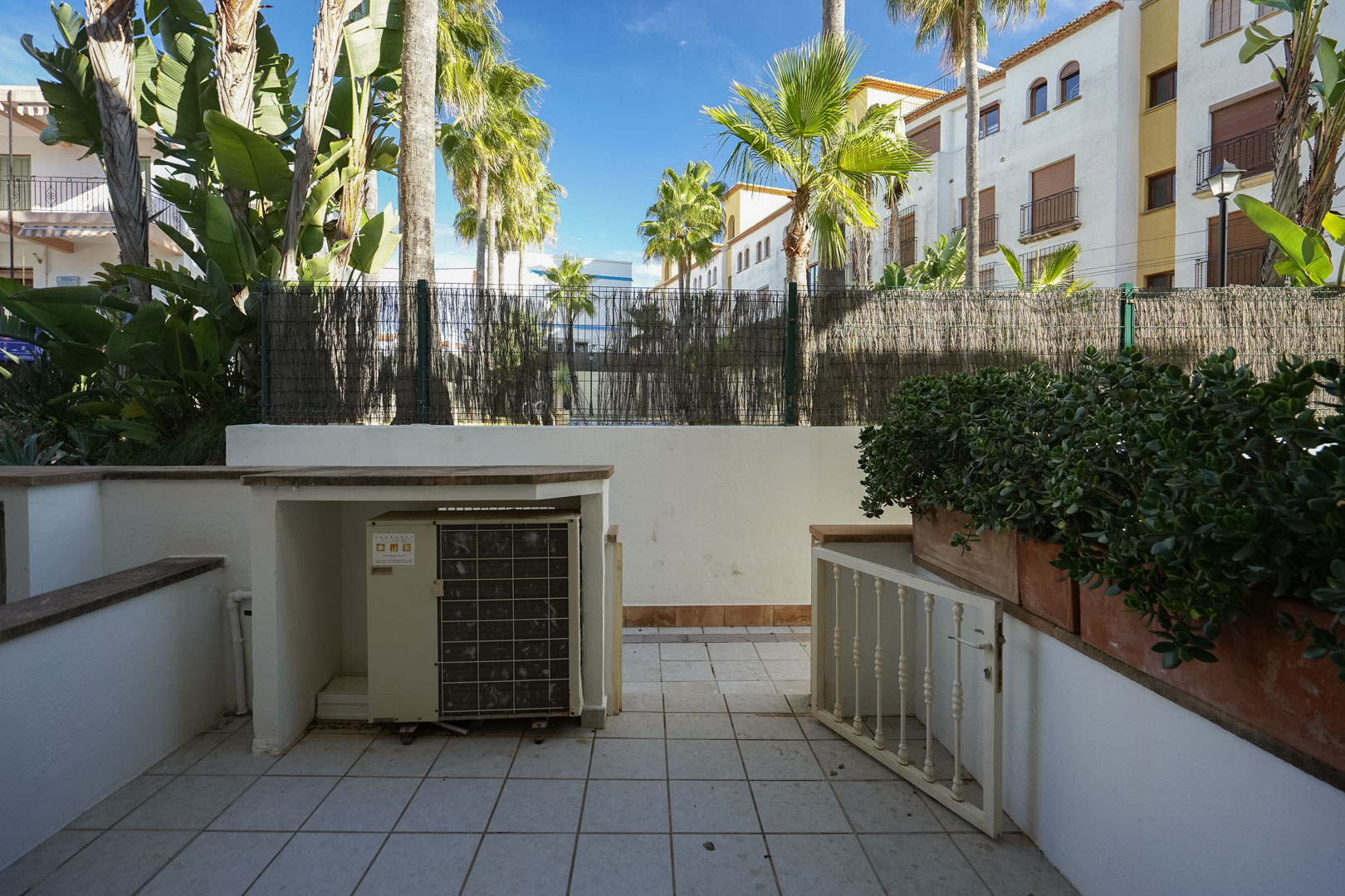 Terrasse privée avec revêtement en céramique, jardinières intégrées et espace pour climatisation, entourée de palmiers et de végétation tropicale.