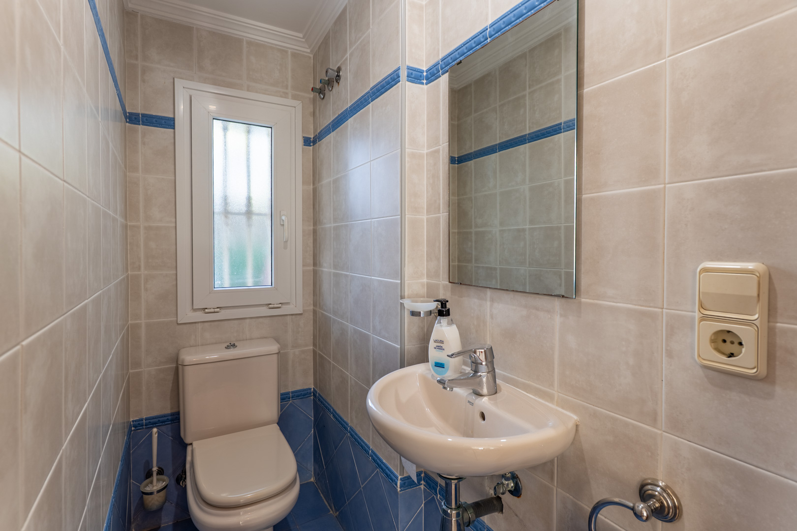 Toilettes invités avec carrelage mural beige et bleu, équipées d'un lavabo compact, d'un miroir rectangulaire et d'une fenêtre oscillo-battante.