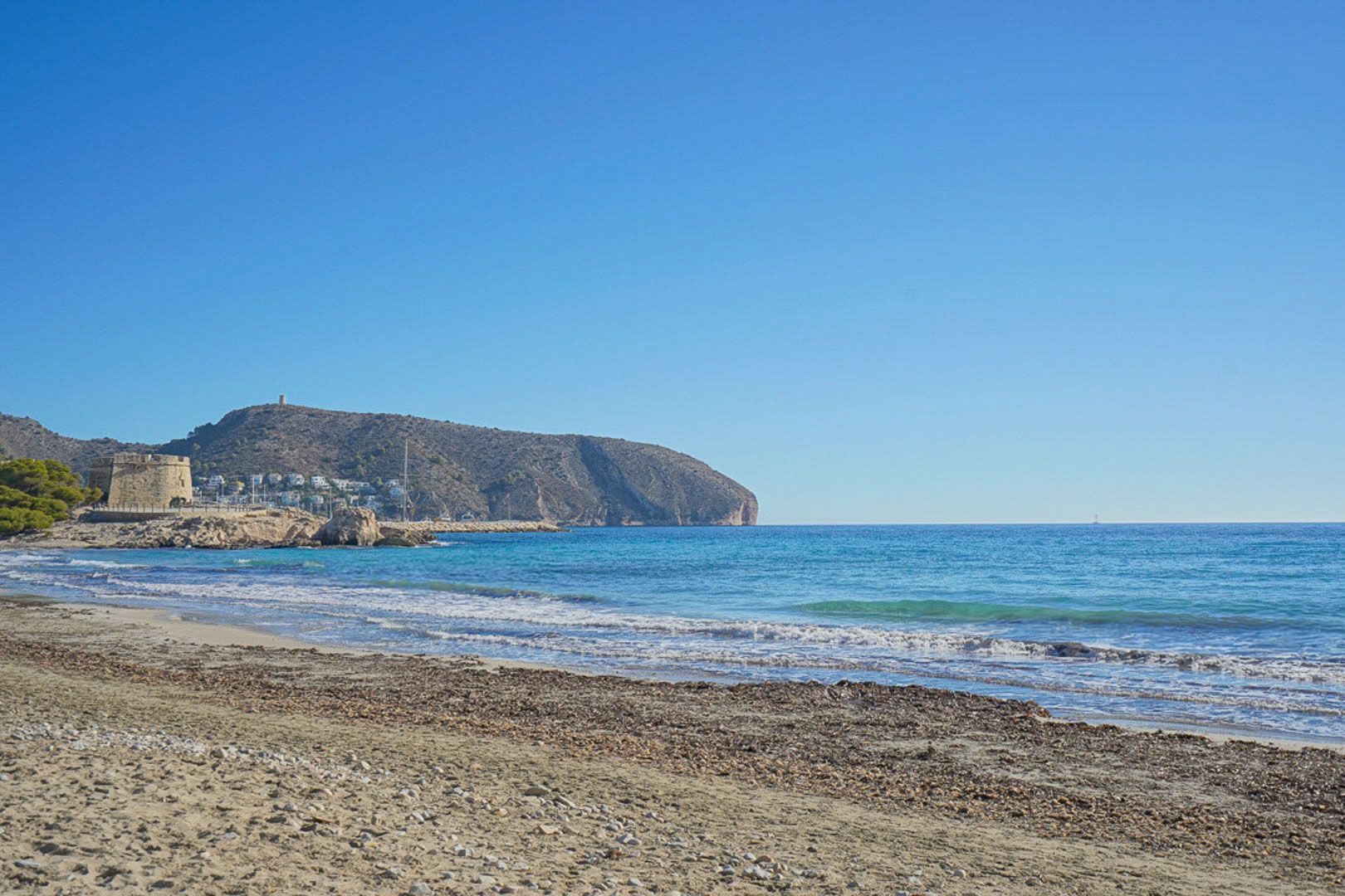 Vue de la plage de Moraira avec sa tour historique et la mer Méditerranée sous un ciel bleu dégagé.