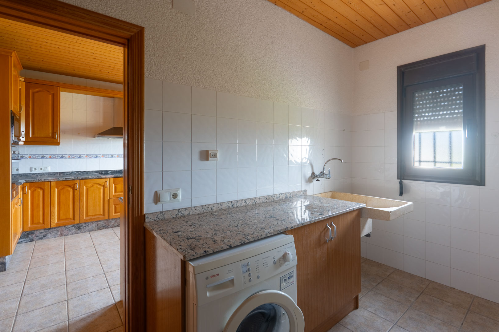 Buanderie équipée d'un plan de travail en granit, d'un lave-linge Bosch et d'un évier en pierre. Plafond en bois et murs carrelés, située à côté de la cuisine.