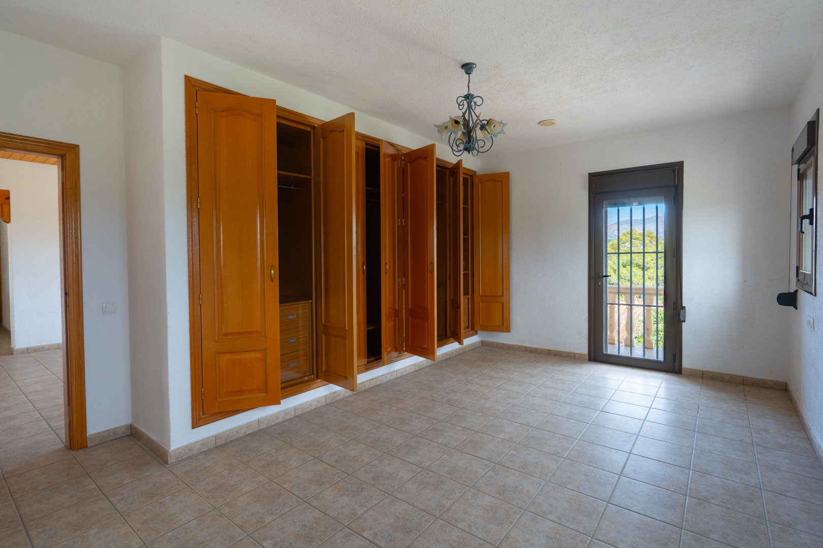 Chambre spacieuse avec une grande armoire encastrée en bois, un sol en carrelage et une porte vitrée ouvrant sur un balcon avec vue sur la nature environnante.