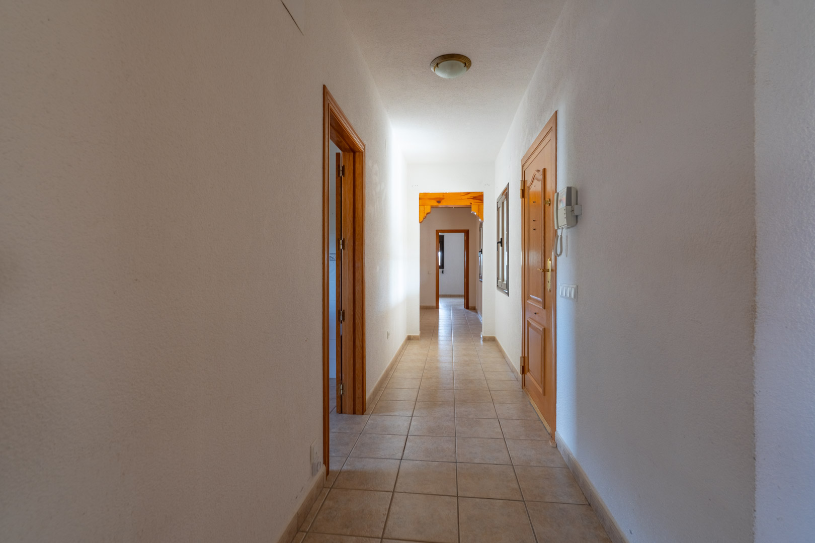 Couloir longitudinal avec sol en grès cérame, murs blancs et portes en bois clair. L'espace dispose d'un interphone et d'une arche décorative en bois massif au fond.