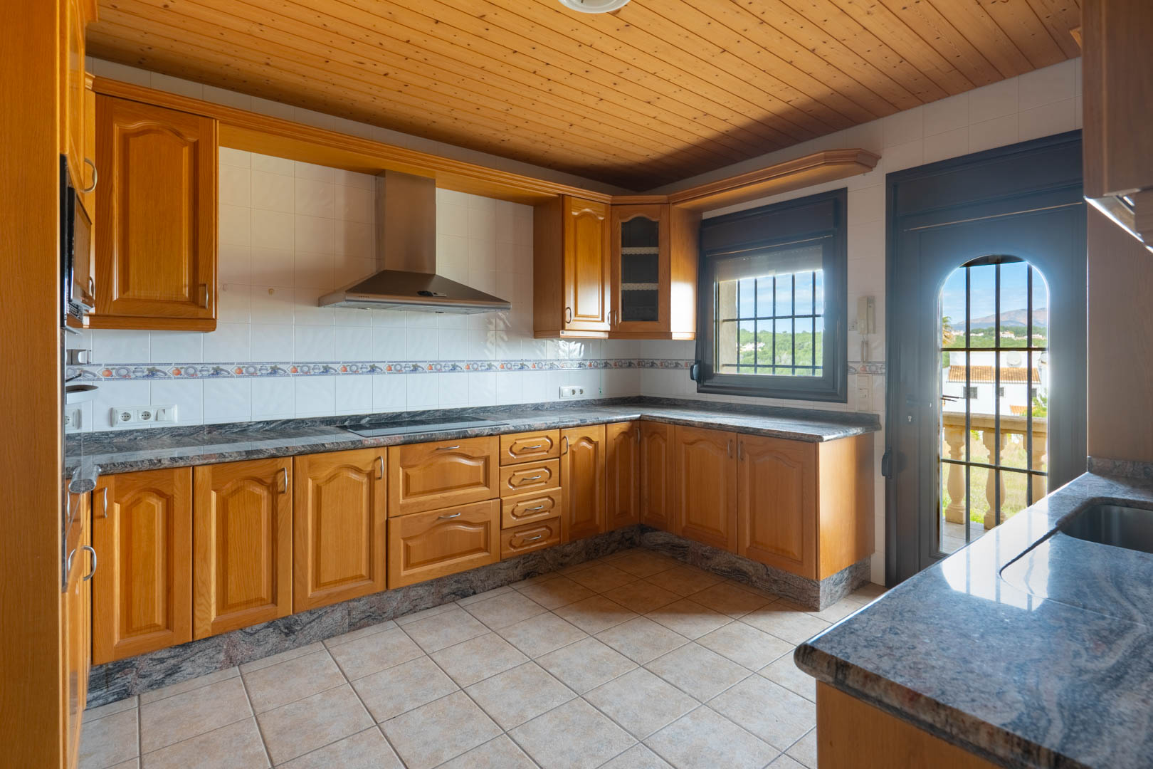 Cuisine avec meubles en bois, plans de travail en granit et plafond boisé. Équipée d'une hotte en inox et d'un accès vers une terrasse offrant une vue sur l'extérieur.