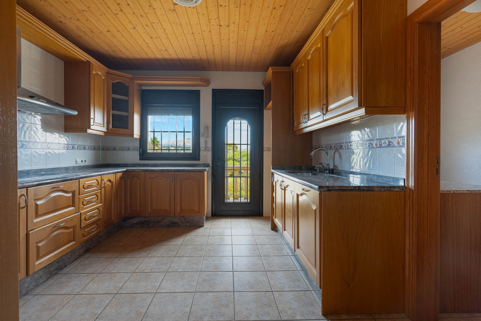 Cuisine avec placards en bois, plans de travail en granit et plafond lambrissé. Elle dispose d'un sol carrelé et d'un accès extérieur vers un balcon avec vue.