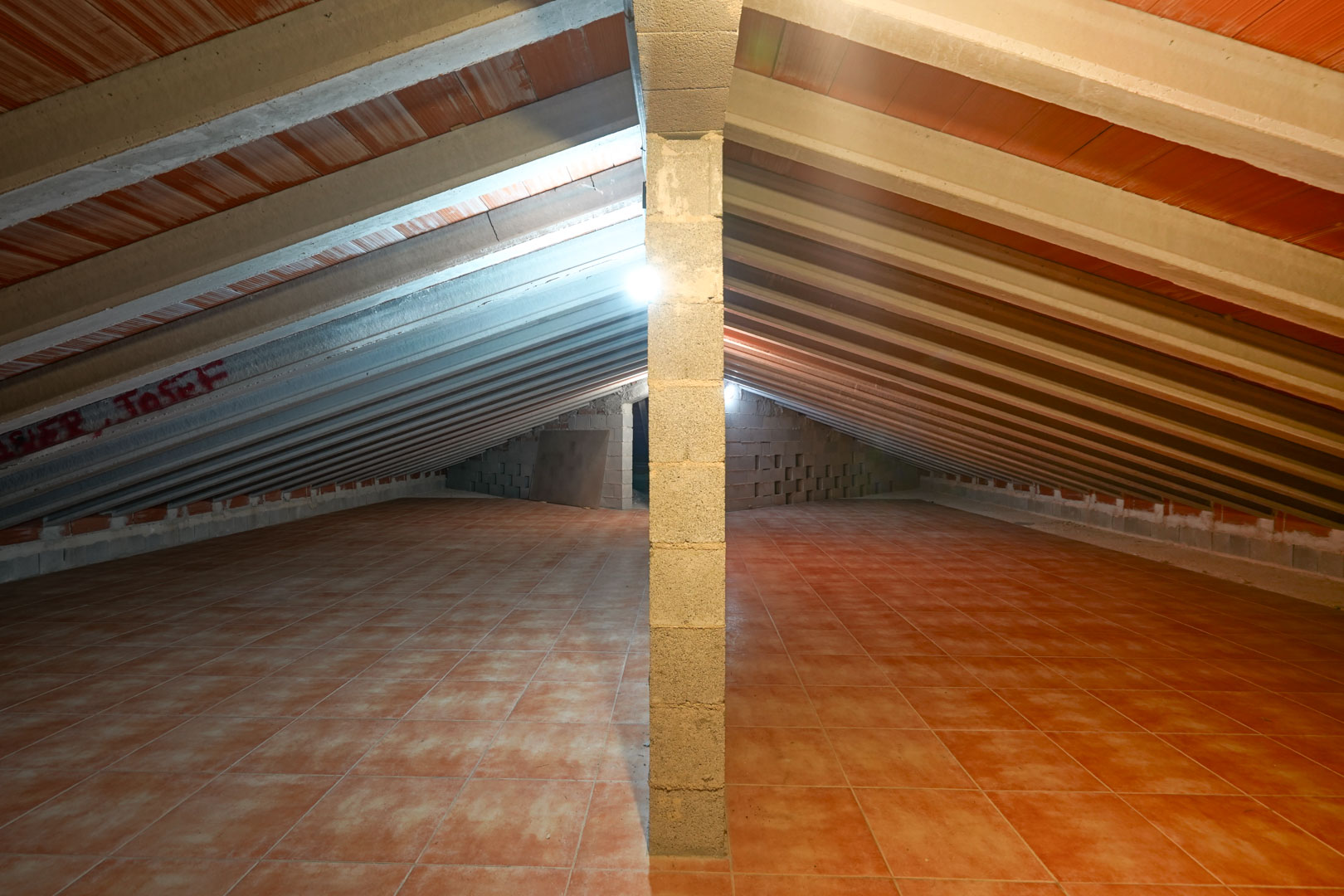 Espace de rangement sous combles avec plafond incliné, poutres en béton apparentes, entrevous en terre cuite et sol carrelé.