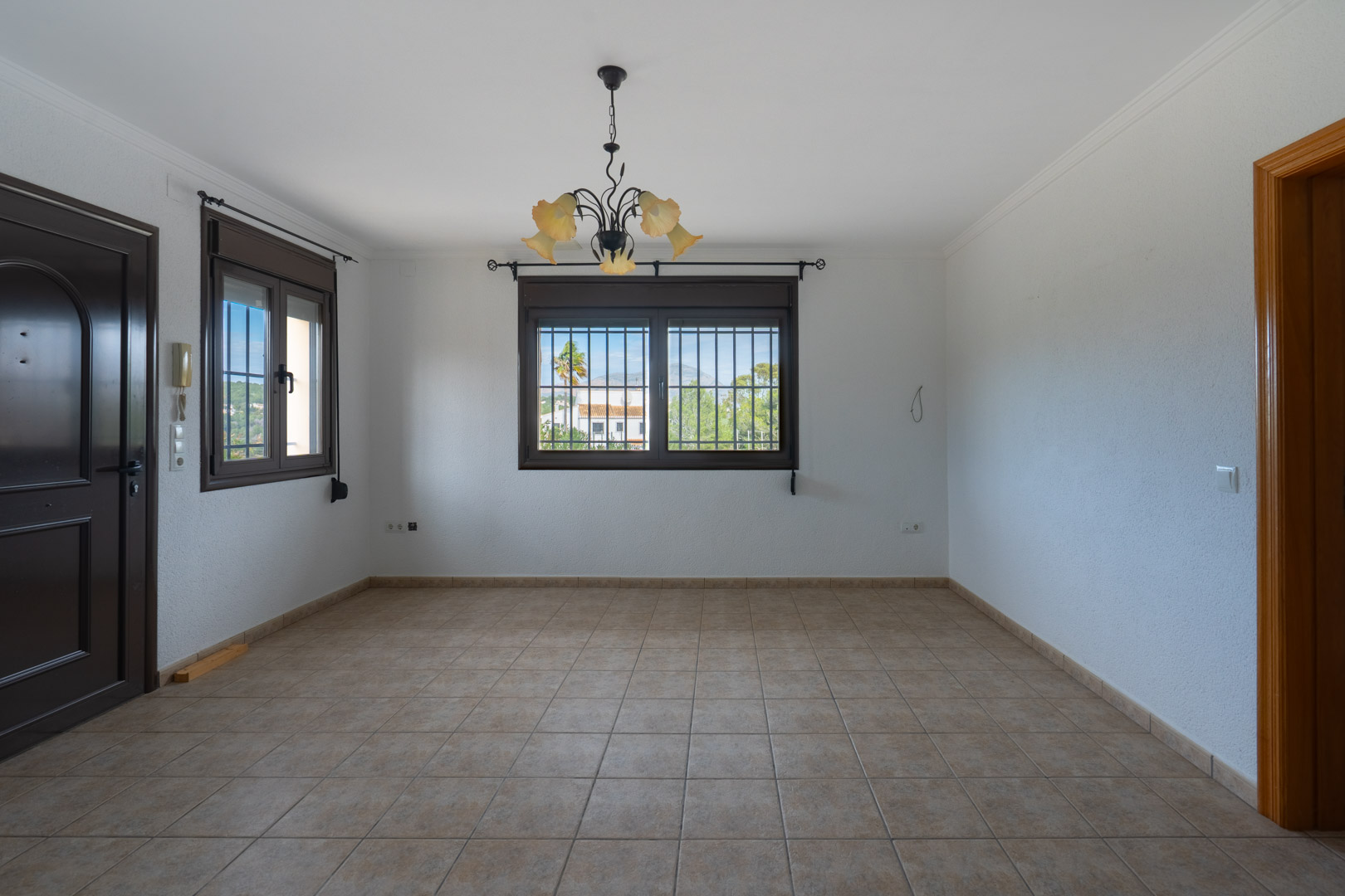 Espace vide avec carrelage beige, fenêtres à cadres sombres munies de grilles de sécurité et lustre en fer forgé aux abat-jours floraux.
