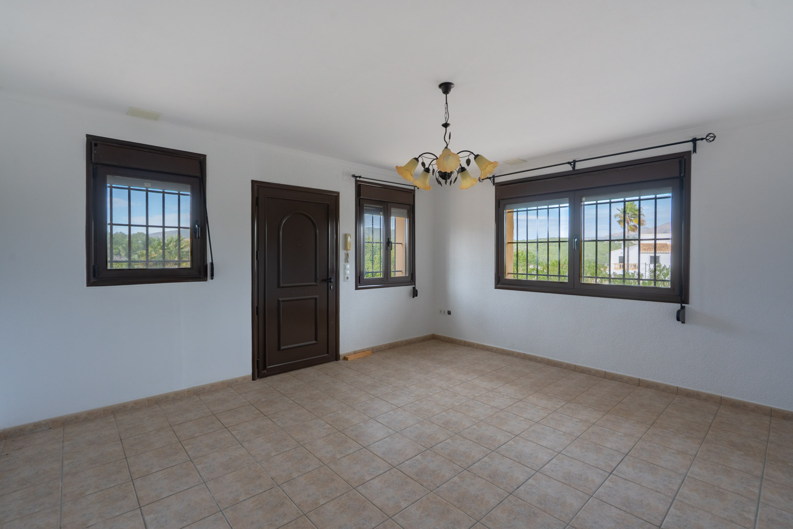 Espace vide avec sol en grès cérame beige, menuiseries brunes avec grilles de protection et lustre floral. Les fenêtres donnent sur un paysage verdoyant.
