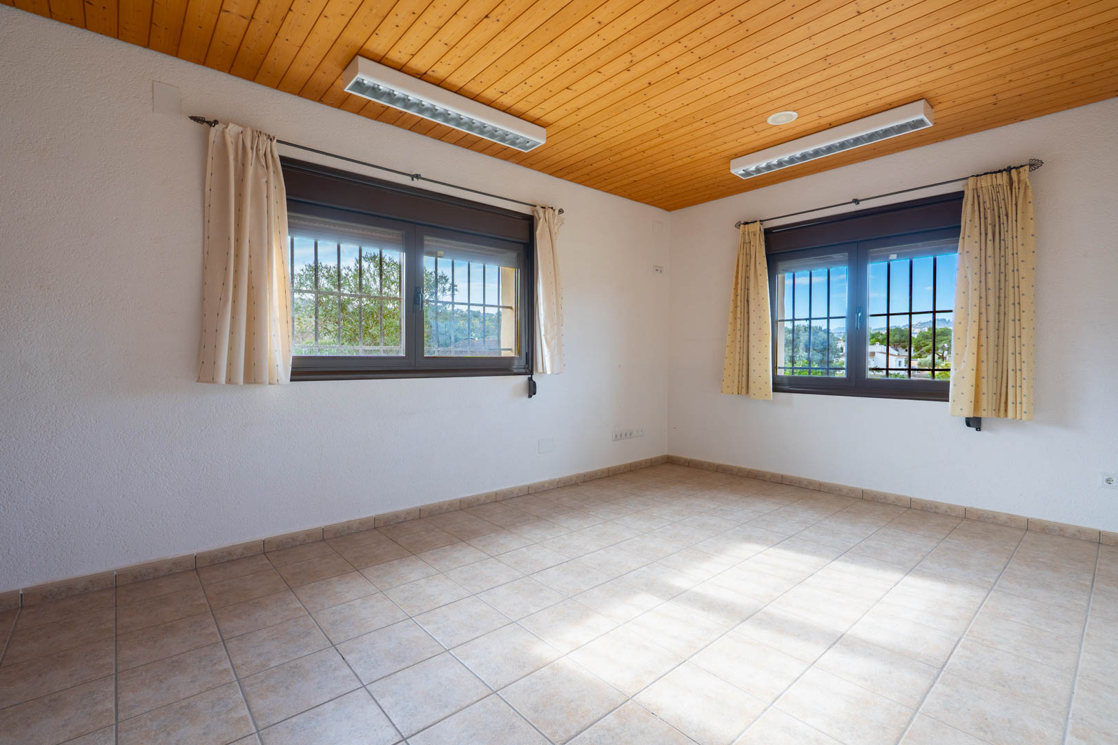 Espace vide doté d'un plafond en lambris de bois, d'un sol en carrelage clair et de deux fenêtres avec grilles de protection extérieures.