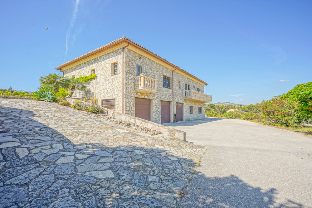 Façade en pierre naturelle avec triple garage, allée pavée et balcons à balustrades, entourée d'un jardin méditerranéen sous un ciel dégagé.