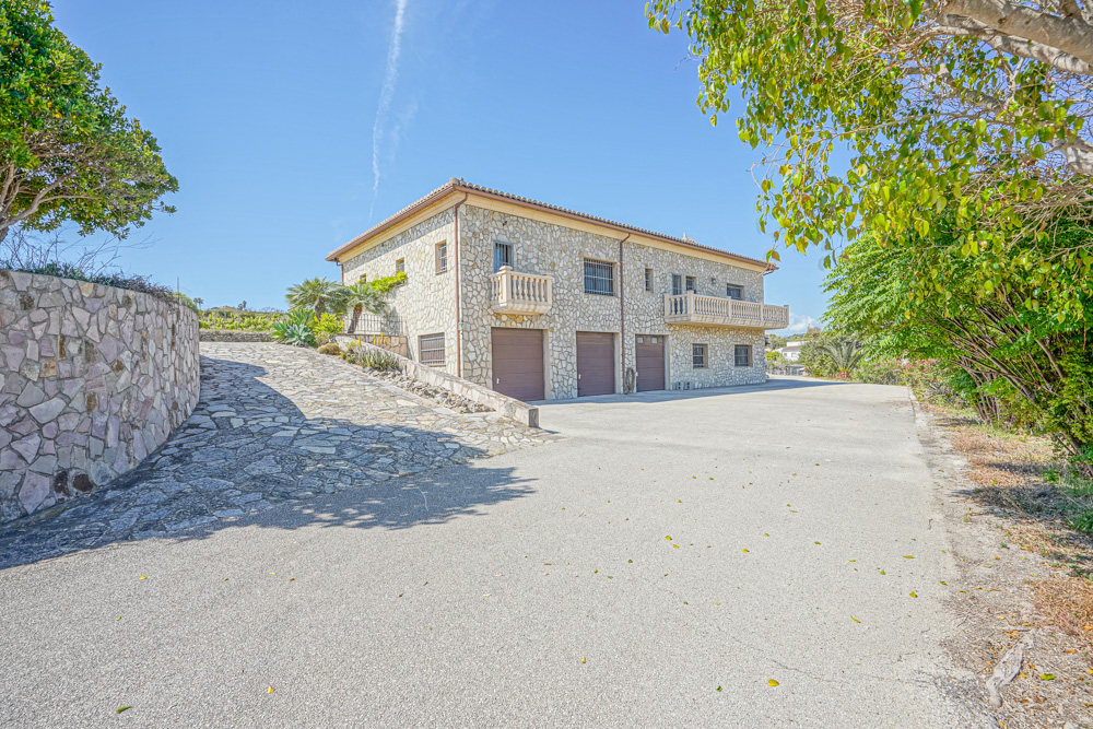 Façade en pierre naturelle avec trois portes de garage, rampe d'accès pavée et balcons à balustrades sous un ciel dégagé.