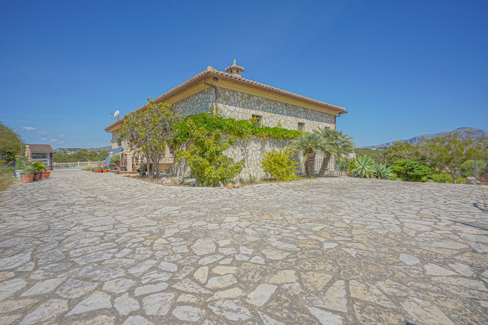 Façade en pierre naturelle avec une vaste allée pavée, jardin méditerranéen comprenant des palmiers et une toiture en tuiles romanes.