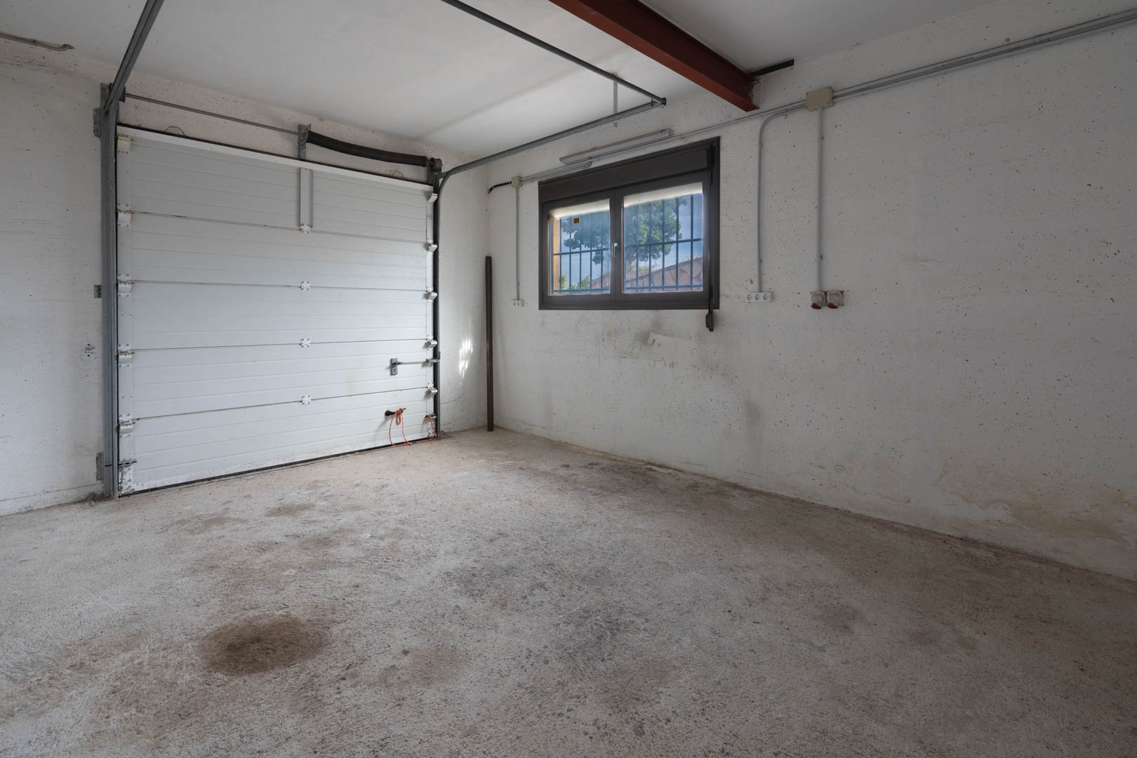 Garage spacieux avec sol en béton, porte sectionnelle blanche et fenêtre sécurisée par des barreaux laissant entrer la lumière naturelle.