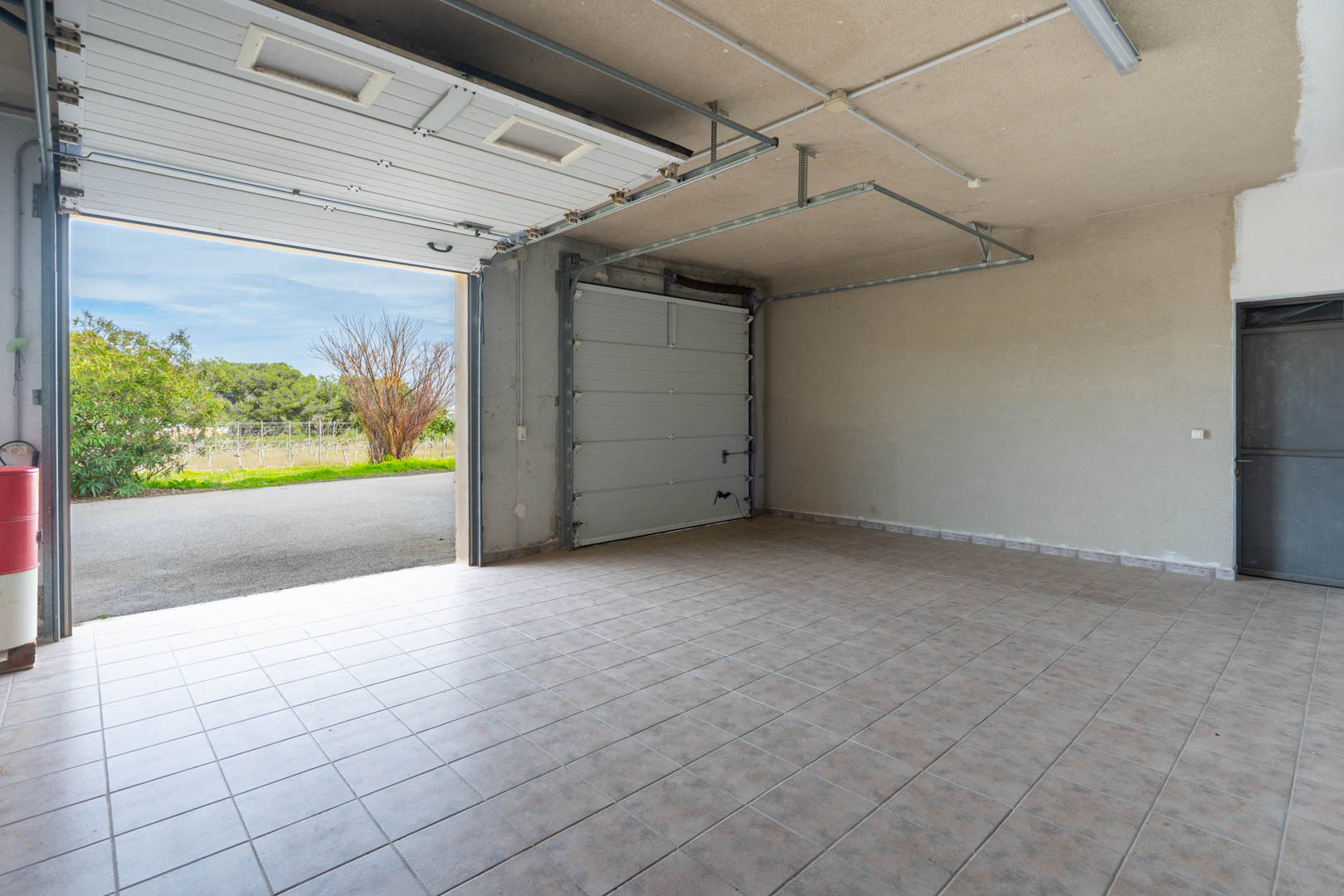 Garage spacieux avec sol en carrelage céramique, doté de deux portes sectionnelles et d'un éclairage au plafond. Accès direct vers l'allée extérieure et le jardin.